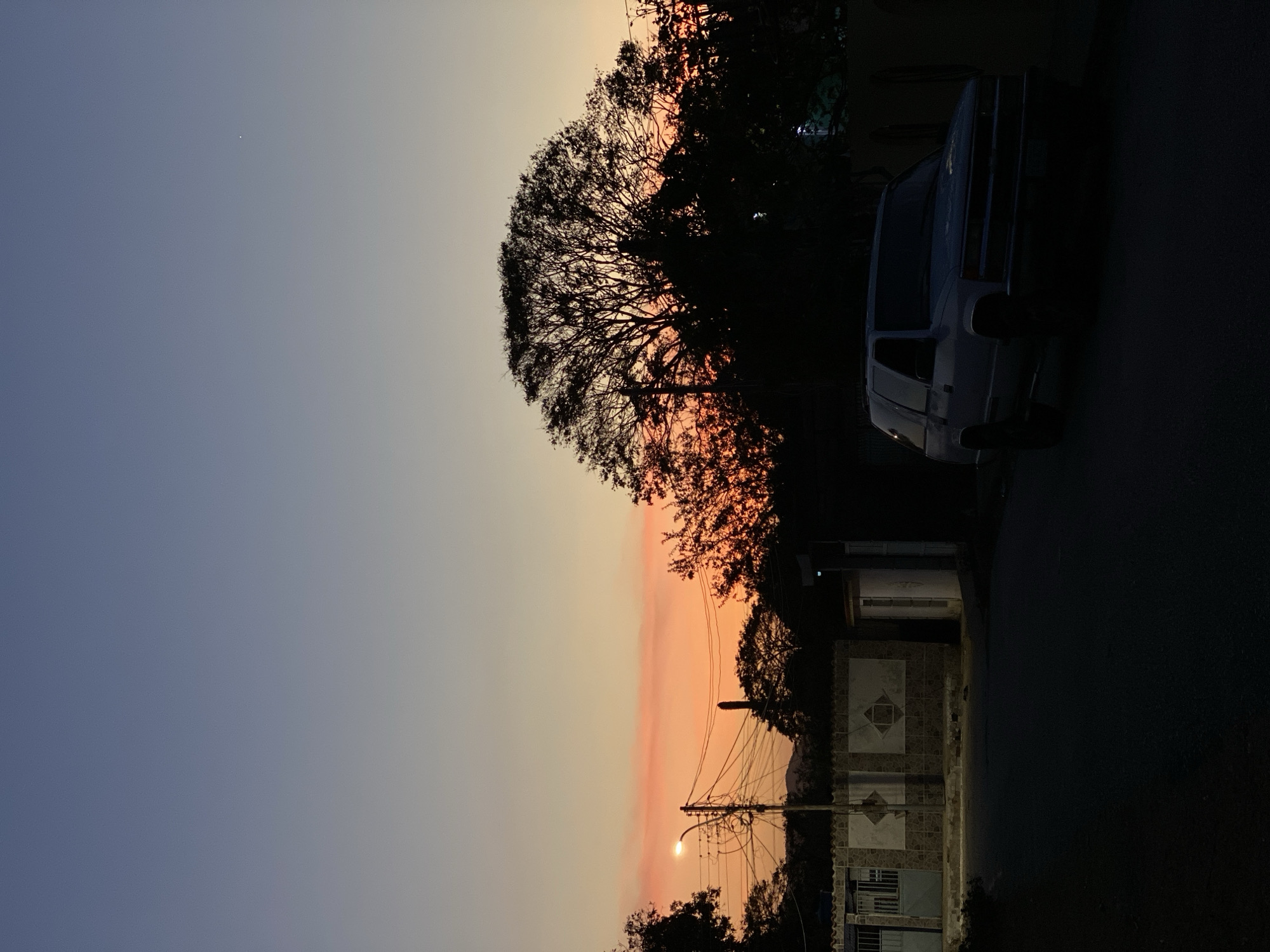 The image depicts a scene at twilight in Juan Griego, Venezuela. The sky transitions from blue to orange, indicating sunset or sunrise. A silhouetted tree dominates the midground with its branches reaching toward the sky. A dark-colored car is parked next to the tree along the road. A streetlamp illuminates the houses. The road occupies a significant portion of the foreground, which is shadowed.