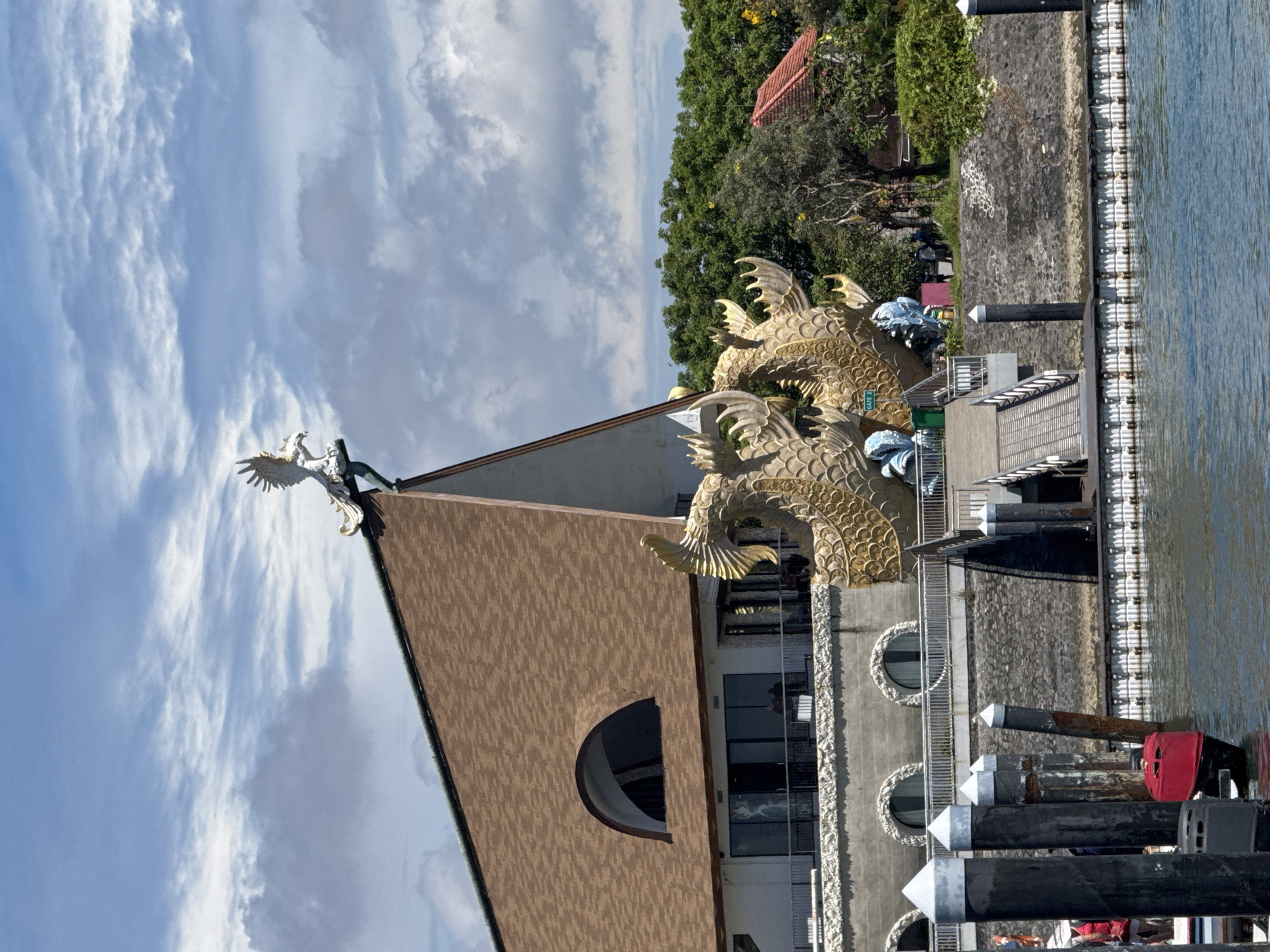 This image, originally in portrait orientation but displayed rotated 90 degrees counter-clockwise, depicts a distinctive building complex situated along a body of water under a bright, partly cloudy sky.
The central subject is a building featuring a prominent A-frame roof with a brown, textured facade. Perched on the apex of the roof is a white statue resembling an eagle or mythical bird with outstretched wings. To the right of the building, a large, ornate golden dragon statue is prominently displayed, intricately detailed with scales and fins. A small green sign with "GATE 2" in white text is visible on the dragon's body. The lower section of the building includes arched windows or openings.
In the foreground, a calm body of blue water, likely a river or canal, stretches out. A stone or concrete embankment separates the building area from the water. Along the water's edge, a white, modular floating dock system is installed. Several dark-colored mooring posts or pylons, some with white pointed caps, rise from the water. A red buoy or fender is also seen near these posts.
The background is dominated by lush green trees and foliage, suggesting a verdant natural setting. A red-tiled roof of another structure is partially visible among the trees. The bright illumination indicates it is daytime. The sky above features scattered white, wispy clouds against a clear blue expanse. There are no discernible people or specific activities taking place, contributing to a tranquil and serene atmosphere. The architectural style, particularly the dragon statue, aligns with the provided location context of Denpasar, Indonesia.