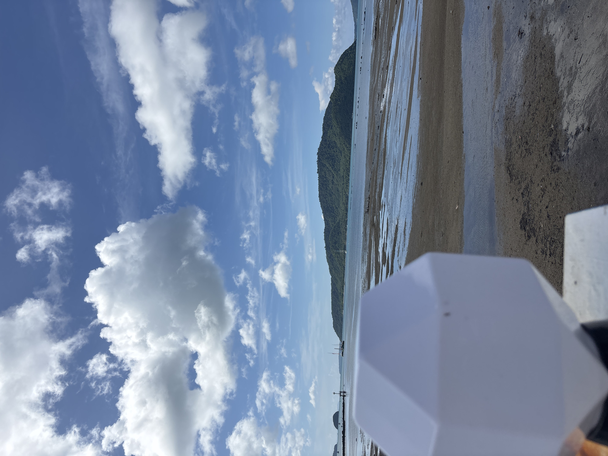 A low tide scene at a beach in Rawai, Thailand, is captured with a foreground element of a white, faceted object, possibly a light fixture or decorative piece, partially obscuring the view. The beach is a mix of wet sand and shallow pools of water reflecting the sky. In the mid-ground, the calm sea stretches towards a large, tree-covered island. Several boats are visible on the water, and a small lighthouse or marker stands on a jetty in the distance. The sky above is a vibrant blue, dotted with fluffy white clouds, suggesting a sunny day. The overall impression is one of a peaceful coastal landscape.