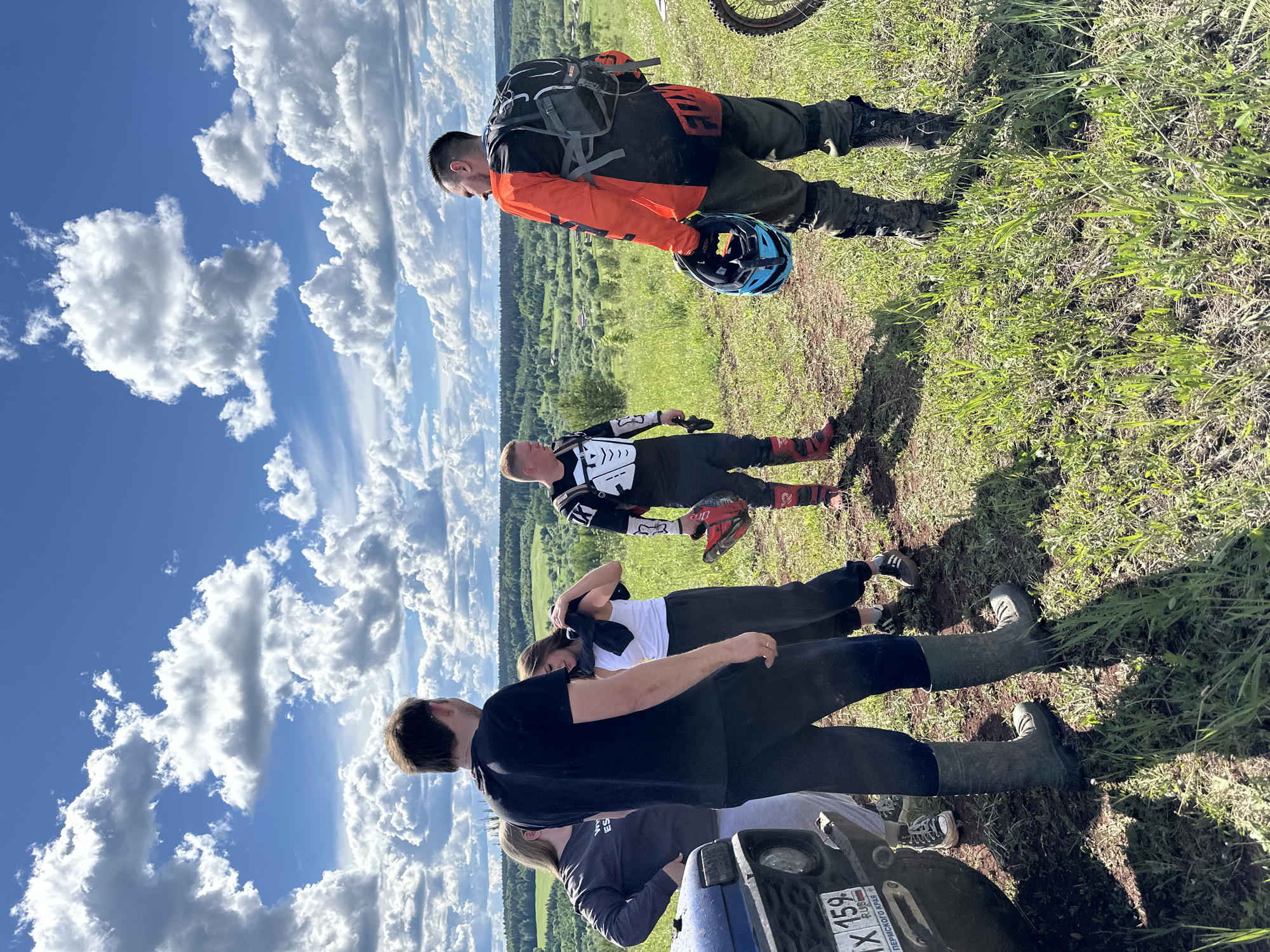 A partly cloudy sky is the backdrop for a group of people standing on a grassy hill. The foreground shows a portion of a blue vehicle with license plate "AX 159," denoting registration in the Perm region of Russia. Four individuals are visible, including two dressed in motocross gear and two in casual clothing. One of the motocross riders holds a helmet. The scene is in Ilyinsky, Russia, indicated by the Cyrillic inscription on the license plate.
