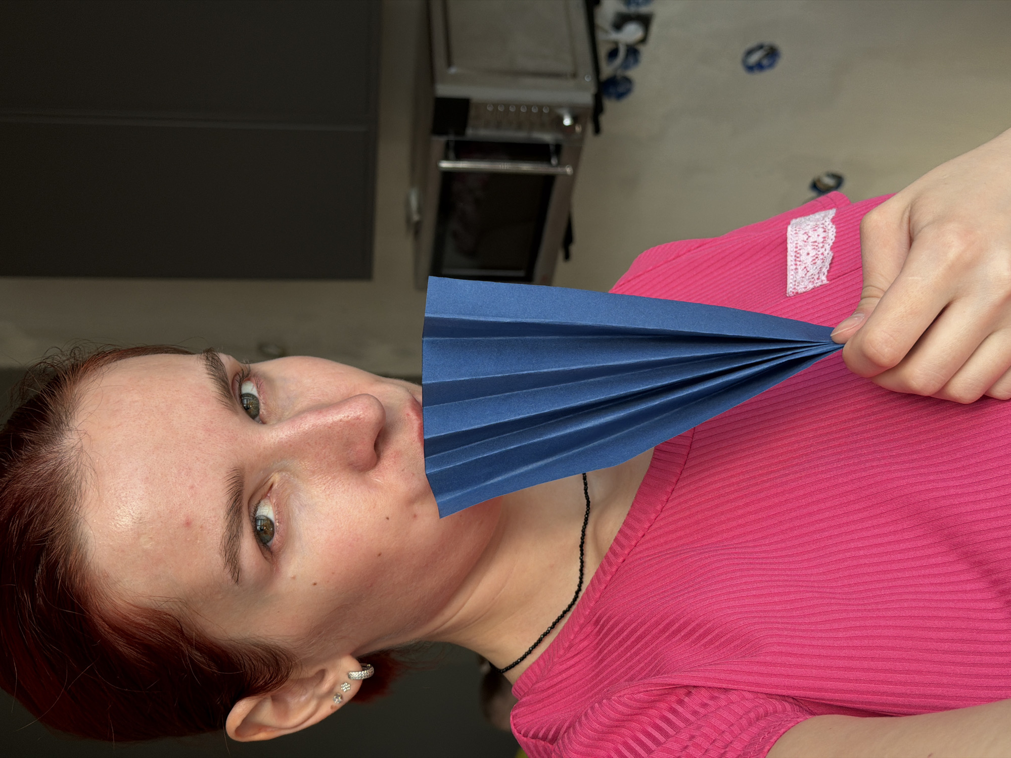 A woman with red hair and fair skin is holding a folded, dark blue paper fan to her face. She is wearing a bright pink, ribbed shirt. The background shows a gray cabinet and a stainless steel oven. The exact location in the city of Chaikovsky, Russia cannot be determined from the image.