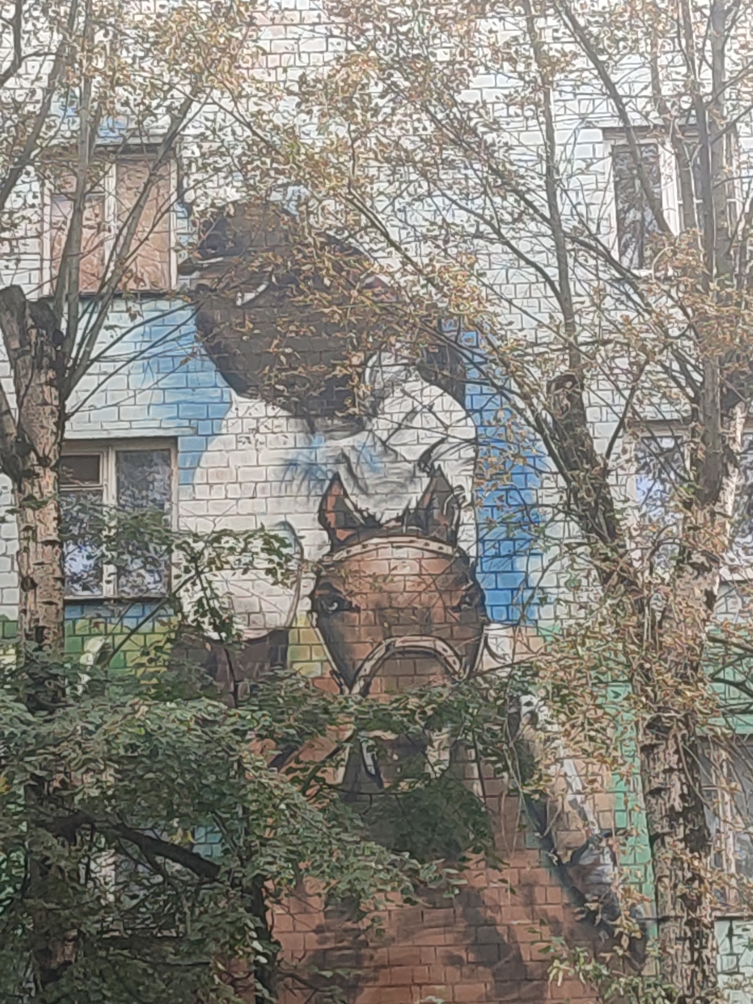 The image depicts a mural on the side of a brick building, partially obscured by trees and foliage. The mural appears to feature a portrait of a horse, with a portion of a human figure visible behind it. The building's facade is made of light-colored bricks, with visible windows. The mural's colors are muted, with shades of brown, blue, and white. The overall scene suggests an urban environment, possibly a residential area in Moscow, Russia, given the architectural style and the presence of trees commonly found in Russian cities. The trees are in a state of autumn foliage, with some leaves having fallen.