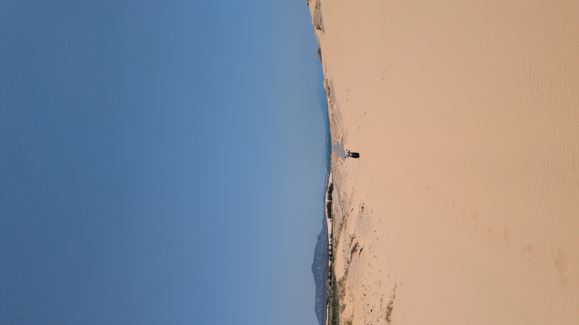 The image depicts a sand dune landscape in Phuoc Dinh, Vietnam, under a clear blue sky. A motorcycle is visible on the sand dune, seemingly parked or stopped. The dunes extend into the distance, with vegetation visible at the base of some dunes. Footprints are evident in the sand.