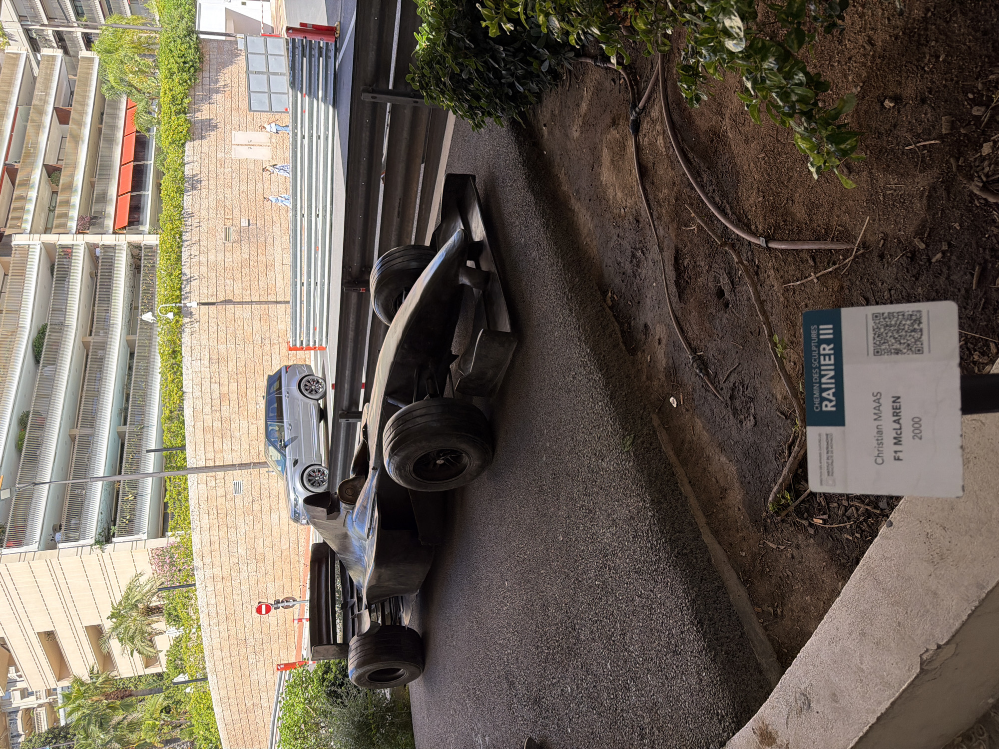 In Monaco, Monaco, a life-sized sculpture of a Formula 1 McLaren car, dated 2000 and created by Christian Maas, is displayed on an asphalt surface. The sculpture is dark in color and positioned prominently on what appears to be part of a street circuit, indicated by the presence of safety barriers and a race track. Behind the sculpture, a silver Range Rover SUV is seen driving on the road. Further in the background, a multi-story building with balconies, lush green vegetation, and palm trees is visible. A sign with text and a QR code is placed in front of the sculpture, providing information about the artwork and its context within "CHEMIN DES SCULPTURES RAINIER III." The lighting suggests it is daytime, with sunlight casting shadows.