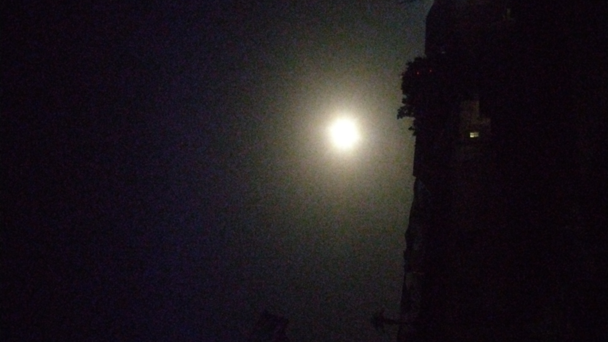 The image is a night shot, seemingly in Jos, Nigeria, featuring a bright light source, presumably the moon, partially obscured by what appears to be a hazy or foggy atmosphere. To the right, a dark, vertical structure is visible, possibly a building or a large tree, with a single illuminated window on the structure. The rest of the image is dominated by darkness, with minimal detail discernible in the shadows. The overall impression is one of a dimly lit, atmospheric scene.