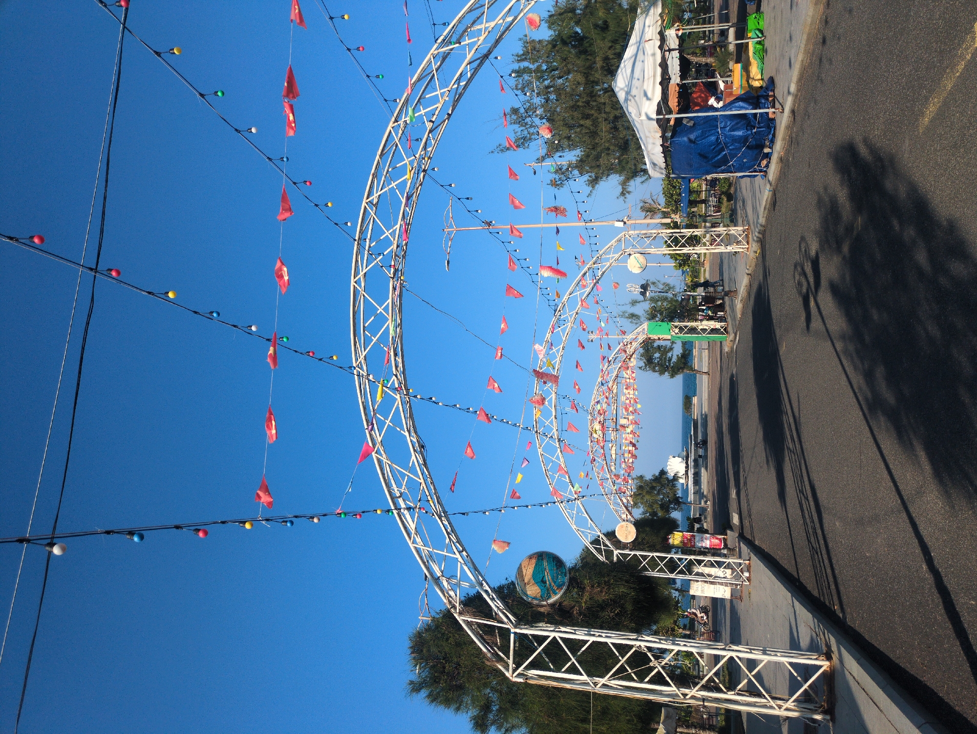 The image shows a street decoration in Tuy Hòa, Vietnam. Arches of white metal framework are decorated with small red flags and strings of colorful lights. The structures line the edge of a paved road. There are vendor tents on the right side of the street. The sky is clear and blue.