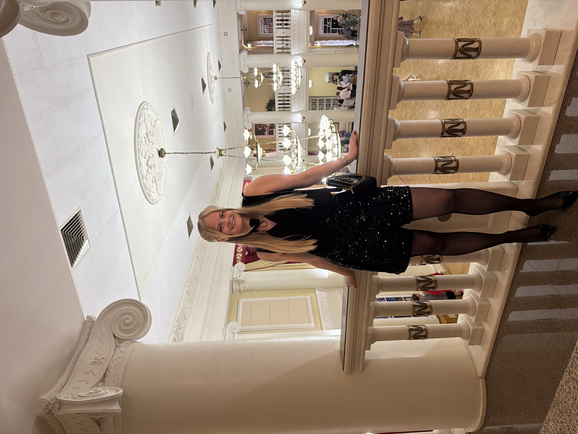 A woman stands on a staircase in the lobby of the State Opera and Ballet Theatre in Izhevsk, Russia. She is positioned with her body oriented perpendicular to the marble balustrade, holding onto it with one hand while looking into the camera. She wears a short, sequined black dress with black stockings and black shoes. She also carries a black purse. The balustrade consists of white marble columns accented with gilded bands depicting leaves. The background reveals a multi-level lobby with chandeliers, columns, and decorations. The ceiling is adorned with ornamental designs. In the background, other people can be seen on different floors of the building. The lobby's color scheme consists primarily of white, cream, and gold.