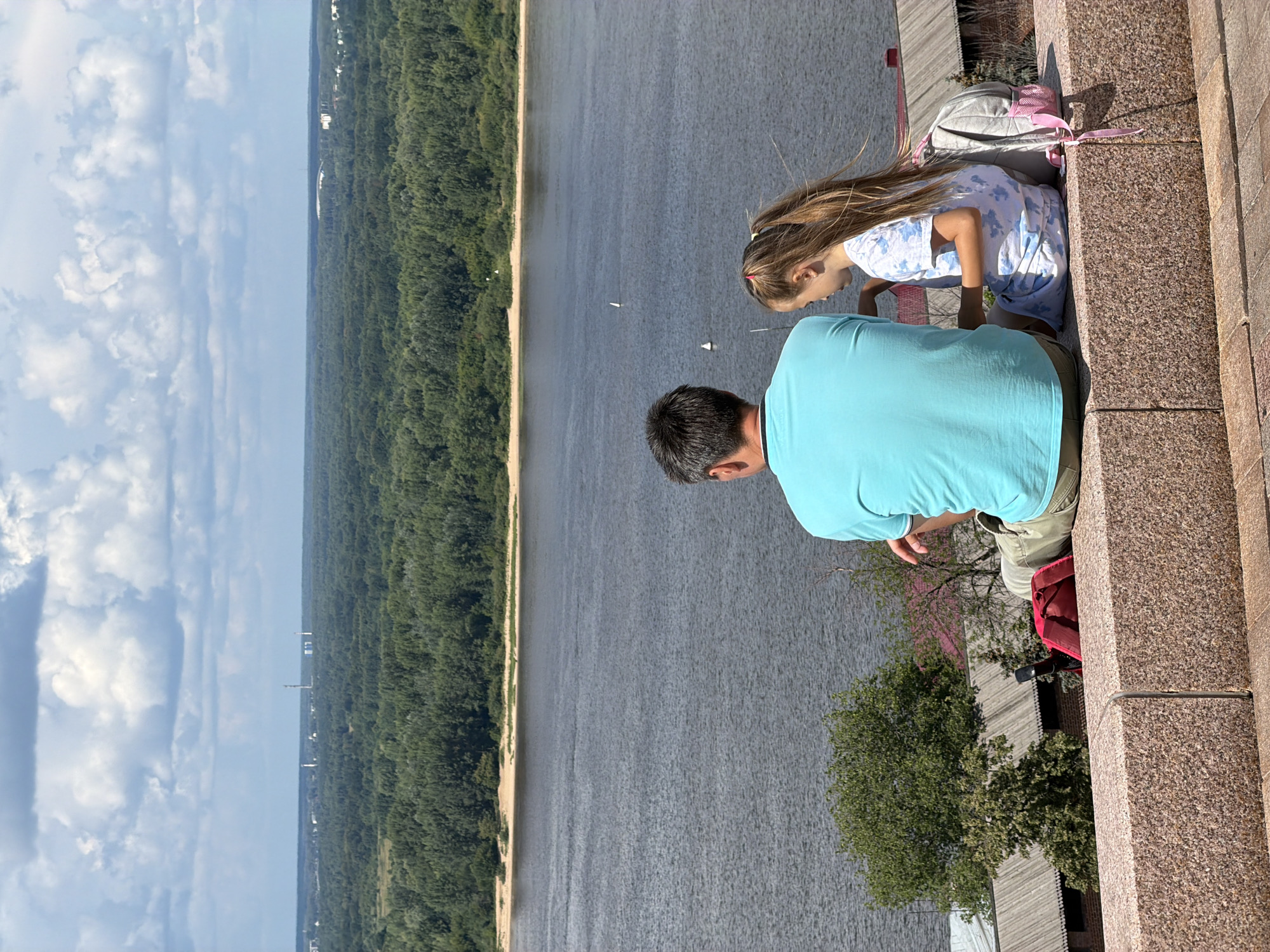 An adult male wearing a turquoise t-shirt and khaki shorts sits on a granite ledge overlooking a wide river. A young female with long blonde hair, wearing a blue and white tie-dye t-shirt and skirt, sits next to him, facing the same direction. She has a grey backpack with pink straps on her right side. In front of them, a red bag is placed on the ledge. The river extends into the distance, bordered by a dense green forest on the opposite bank. A hazy skyline with industrial structures is visible on the horizon under a cloudy blue sky. The scene is likely the Nizhny Novgorod Kremlin, a historical fortress overlooking the confluence of the Oka and Volga rivers, offering panoramic views of the city and surrounding landscape.