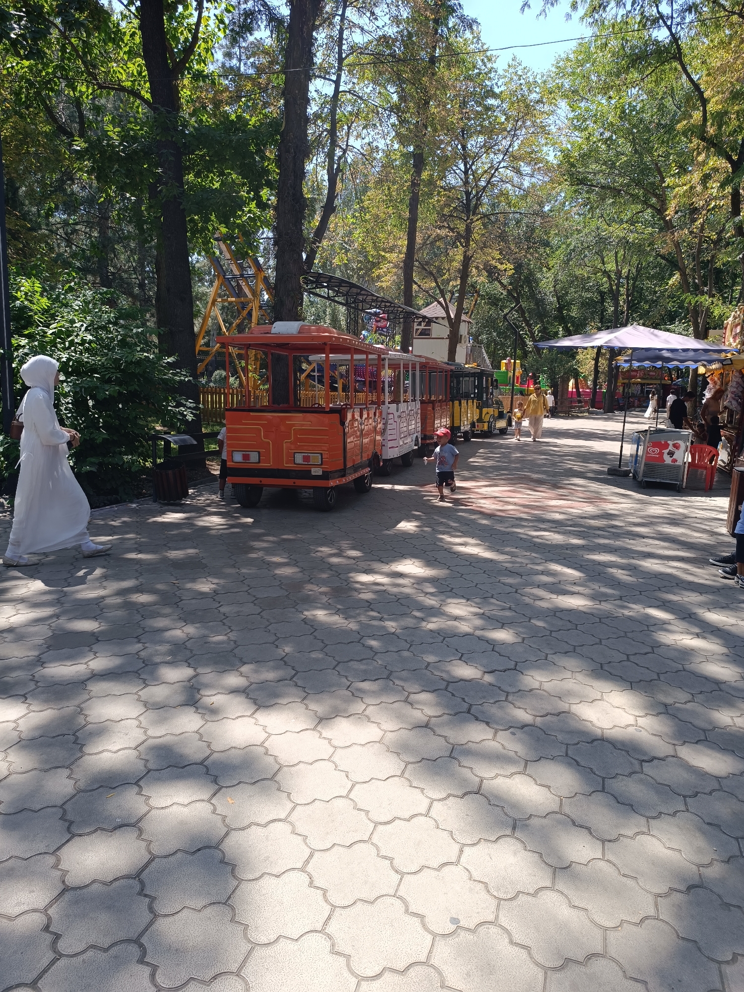 The image depicts a sunny day in what appears to be an amusement park or a public recreation area in Bishkek City, Kyrgyzstan. The foreground is dominated by a paved walkway with a decorative, interlocking stone pattern, shaded by the dappled sunlight filtering through the overhead trees. On the left side of the walkway, a person dressed in a white, hooded garment, possibly a burqa or niqab, is walking away from the camera.
Further into the scene, several amusement park attractions are visible. An orange and white miniature train, likely part of a ride, is parked on tracks. Behind it, other colorful rides and structures, including a yellow Ferris wheel structure and a covered tent-like stall, suggest a lively atmosphere. A few people, including children, can be seen walking along the path or near the attractions. The environment is lush with mature trees providing ample shade, indicating a well-established park. The overall impression is one of a public space designed for leisure and entertainment.