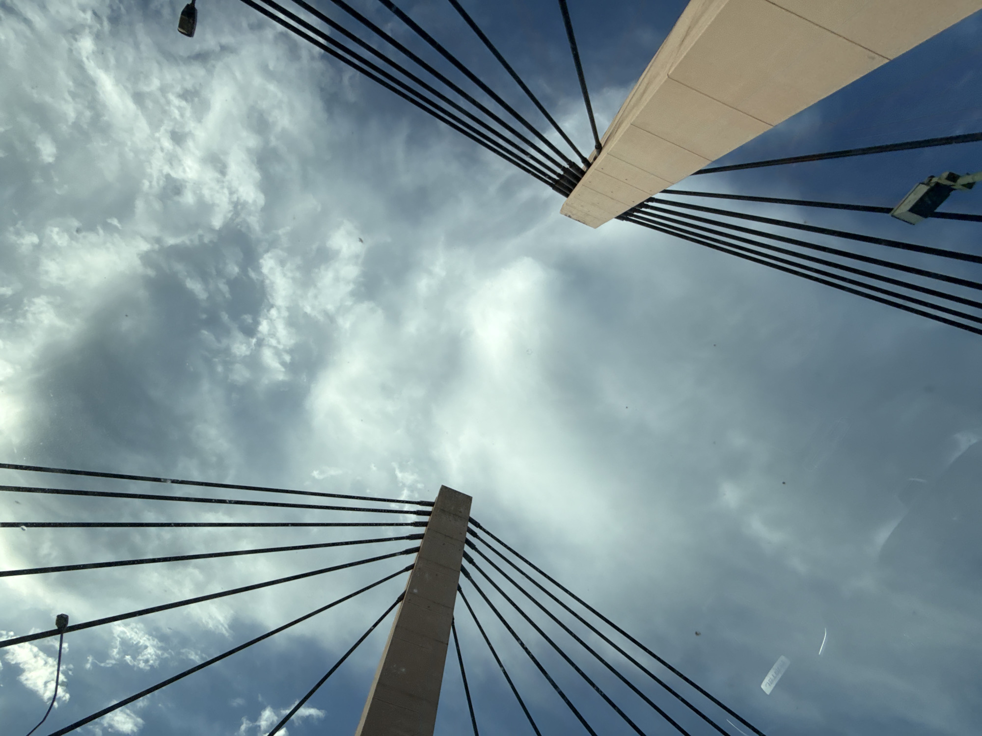 This is a low-angle, upward-facing shot of two pedestrian bridge towers, likely part of the Grunwald Bridge in Wrocław, Poland, viewed through a reflective surface, possibly a car windshield. The sky is a mix of blue and cloudy, with dramatic light and shadow. The bridge towers are a light beige color and are connected to the ground by numerous dark cables that fan out overhead. One tower is on the left side of the frame, angled from bottom left to top right, and the other is on the right side, angled from bottom right to top left. Light fixtures are visible on the cables of the right tower. The reflective surface introduces distortions, smudges, and what appear to be light leaks or reflections from inside a vehicle.