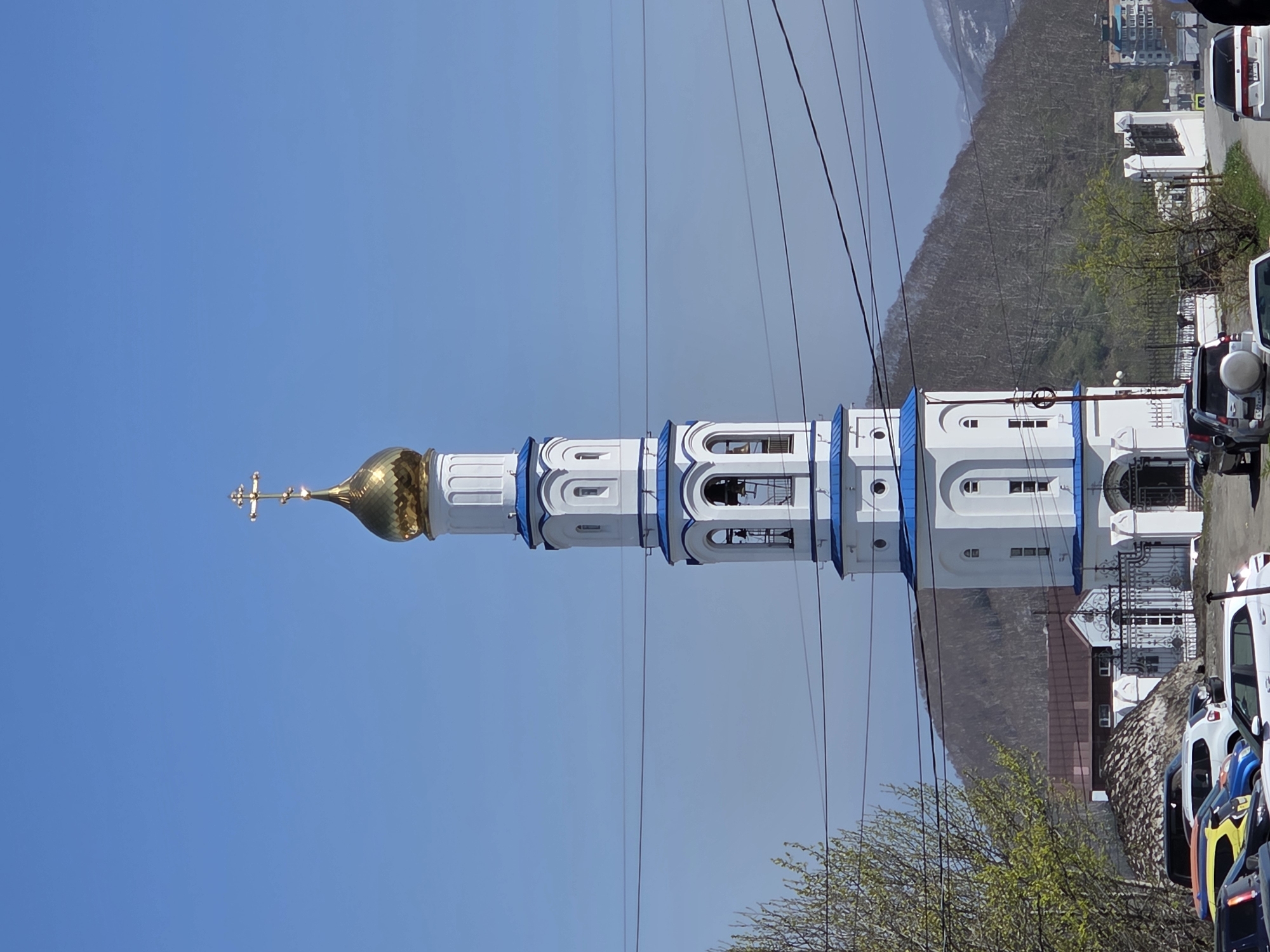 The image displays a tall, white bell tower with blue architectural accents, capped by a golden onion dome and an Orthodox cross, standing against a clear blue sky. The tower is the central and dominant subject. In the background, a range of mountains covered in dense, mostly bare trees, with patches of new green foliage, stretches across the mid-ground.
Several dark, thin power lines or wires traverse the upper part of the image, casting faint shadows against the sky. In the foreground and mid-ground on the right side of the image, a paved road or street is visible with several parked vehicles, including a white van and a silver SUV. Small trees with light green leaves are scattered throughout the lower right portion of the scene. Parts of other buildings, including what appears to be a decorative metal gate, are visible near the base of the bell tower and behind the cars.
The scene depicts a bright, clear daytime, likely morning or early afternoon, with sunny weather. No specific activity or event is taking place; it is a static architectural and landscape shot. The architectural style of the bell tower, particularly the onion dome and cross, suggests an Eastern Orthodox church or monastery, consistent with the given location context of Petropavlovsk-Kamchatsky, Russia. No visible text is clearly legible on any of the objects, including vehicle license plates.