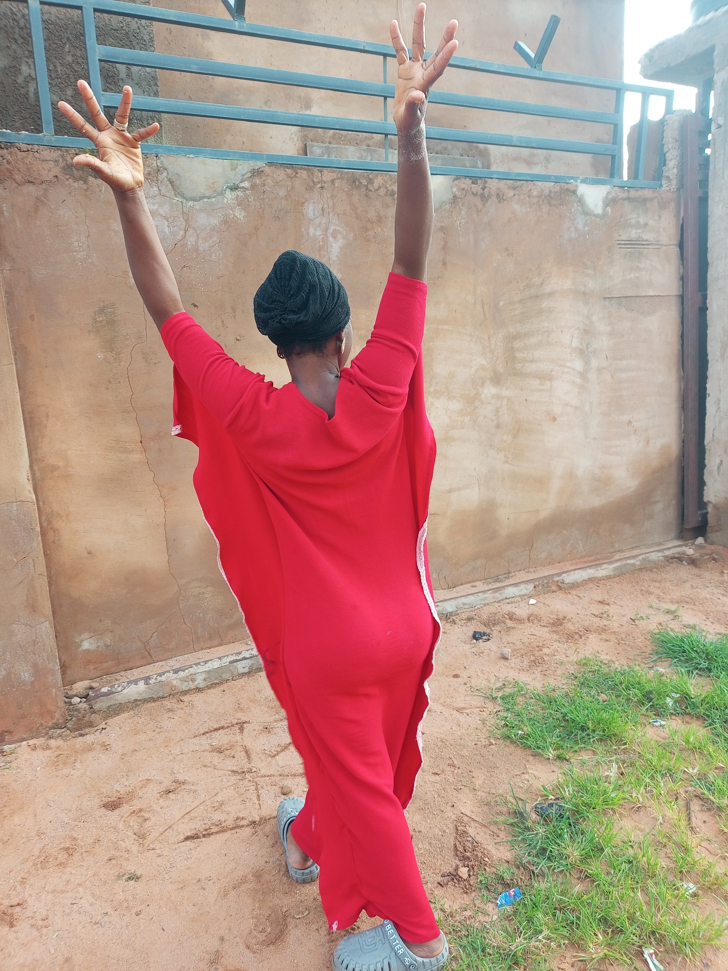 A person, viewed from behind, stands with arms raised and spread wide, wearing a bright red garment and a black head covering. The individual is standing on a dirt and sand surface, with sparse patches of green grass to the right. A beige, rough-textured wall forms the backdrop, with horizontal metal bars running across the upper portion. The scene appears to be outdoors, possibly in a residential area. The person's feet are clad in gray sandals. The image is taken at eye level. The lighting is bright and natural, suggesting daylight.