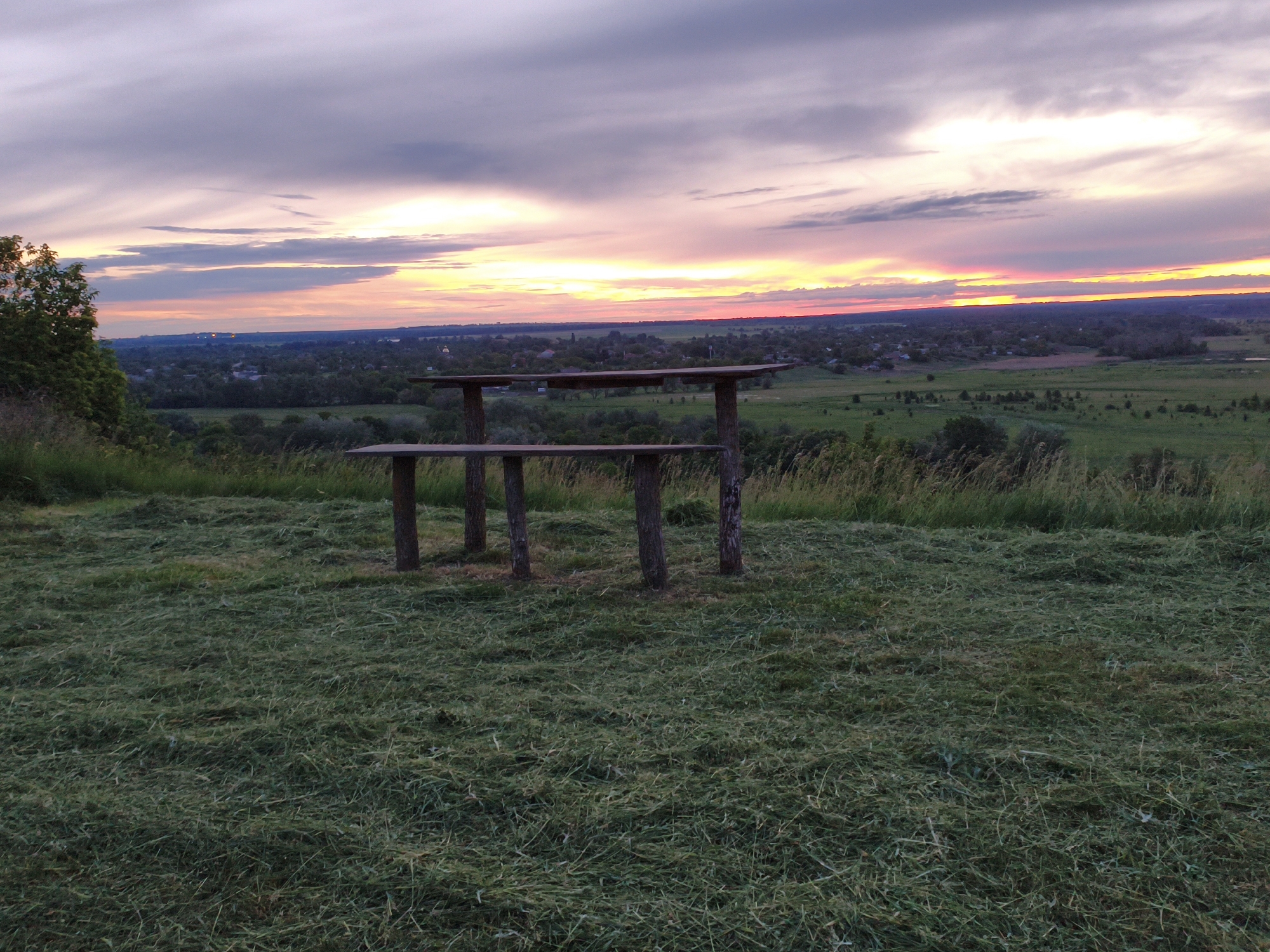 The image depicts an outdoor scene at sunset. A wooden table with two tiers stands on a grassy hill overlooking a landscape of fields, trees, and a distant town. The sky is filled with colorful clouds, ranging from purple and pink to orange and yellow near the horizon. The grass in the foreground appears to have been recently cut. The location is in an unknown city.