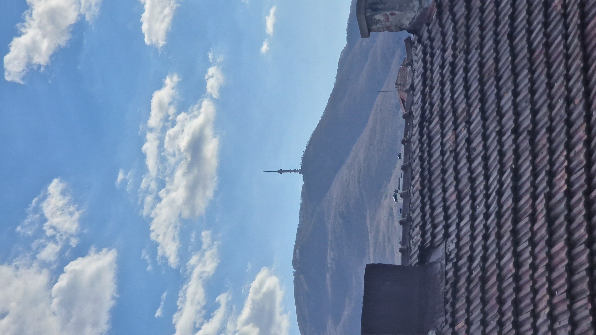 The image shows a view of Sofia, Bulgaria, with a clear blue sky scattered with white clouds on the left side. A mountain range is visible in the center, with the Sofia TV Tower standing atop the mountain. To the right, a roof of red clay tiles dominates the foreground, with a building structure partly visible at the top edge. The orientation suggests the image was taken from an elevated position, possibly from a building window or balcony.