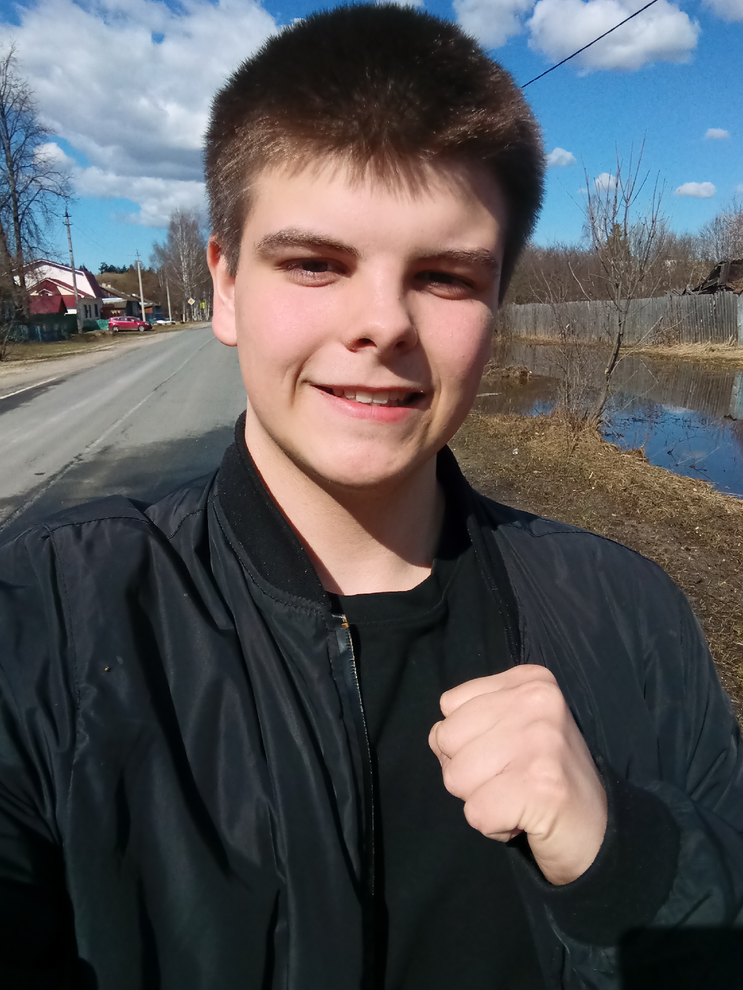 A young adult male is posing for a selfie. He is wearing a black bomber jacket over a black t-shirt. He has short brown hair and is smiling slightly. His left hand is clenched into a fist.
The background shows a rural scene. There is a paved road to the left and a body of water with vegetation on the right. A wooden fence stands between the water and the road. In the distance, there are houses and trees. The sky is partly cloudy. Based on the description of the location, Красный Маяк, Russia, the photo likely depicts a rural area within or near the city where flooding is possible. The season appears to be early spring, as evidenced by the bare trees and the presence of standing water.