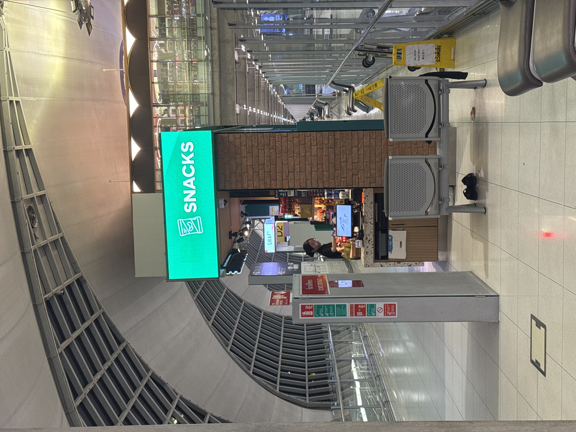 An indoor scene, likely an airport terminal, shows a snack kiosk with a bright green sign displaying "SNACKS" and a graphic of what appears to be a juice box. A female employee wearing glasses and a dark uniform stands behind the counter, interacting with a computer system. The kiosk features a brick facade. In front of the kiosk are two empty, grey chairs with perforated seats. On the floor next to the chairs are a pair of black shoes. Further down the corridor, more empty seating areas are visible, separated by metal railings and glass partitions. A yellow caution sign with black text reading "NO ENTRY STAFF ONLY" and some text in Thai is also present. The overall environment suggests a public transit hub, with modern architecture characterized by curved ceilings and tiled floors. The location is identified as Nong Prue Subdistrict, Thailand.