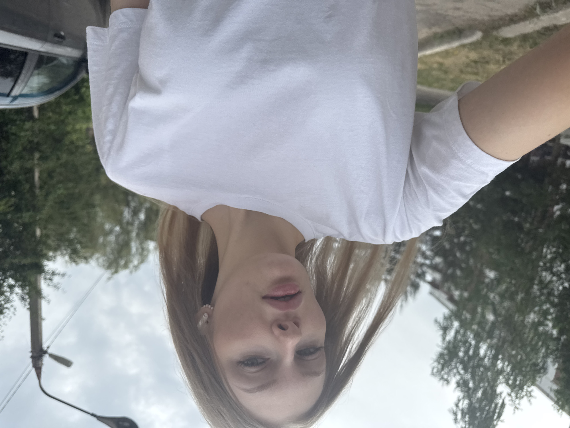 A woman with long blonde hair wears a white t-shirt and pearl earrings while standing outside in Perm, Russia. The background includes trees, a car, a street light, and buildings. The sky is cloudy.