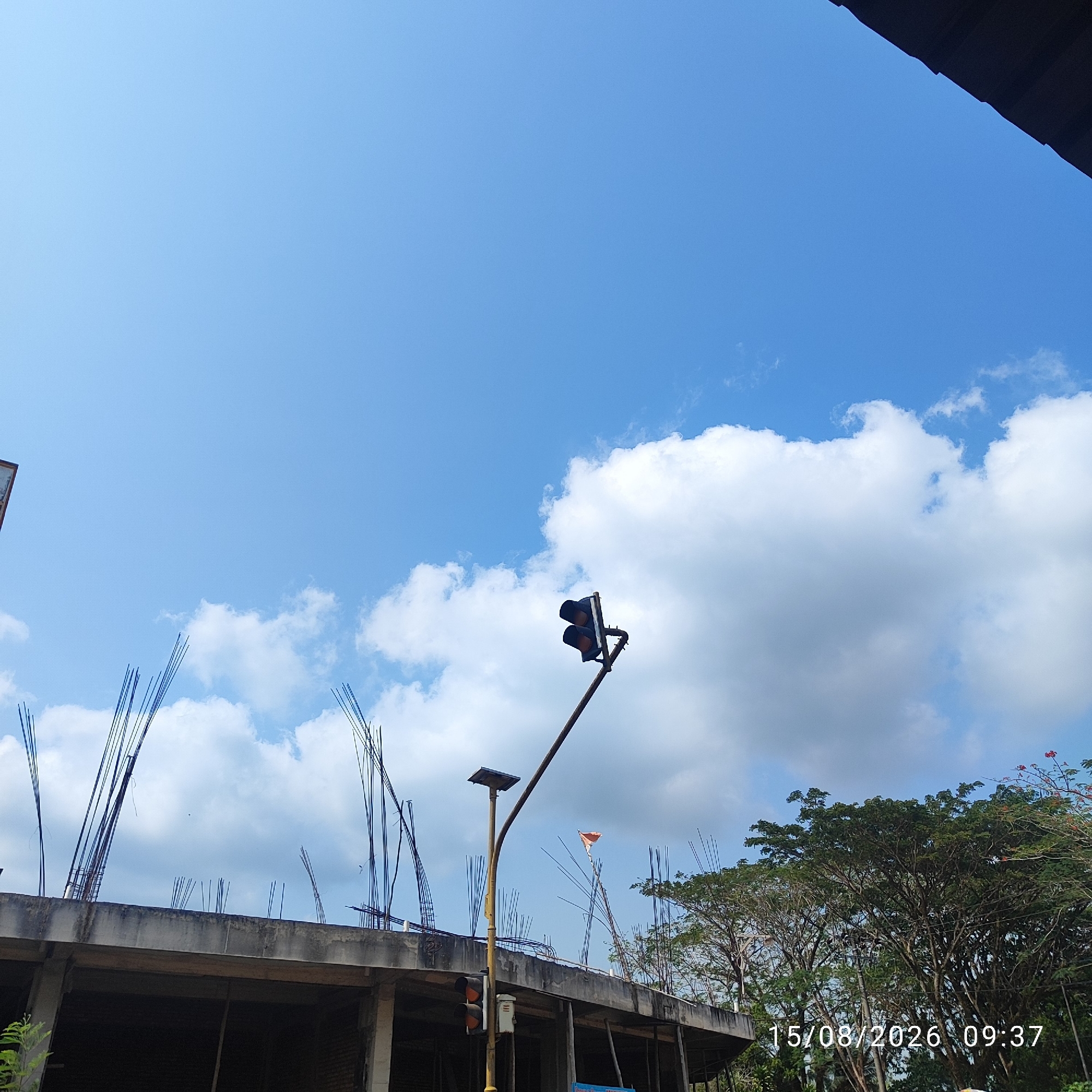 The image depicts an outdoor scene with a bright blue sky filled with scattered white clouds. In the foreground, a partially constructed building with exposed rebar is visible. A traffic light, with its yellow lights illuminated, is mounted on a pole that also supports a small solar panel. A small orange flag is attached to a pole behind the traffic light. To the right of the building, a dense canopy of green trees dominates the middle ground. The lower right corner of the image shows a timestamp: "15/08/2026 09:37". The overall scene suggests an urban or semi-urban environment undergoing construction.