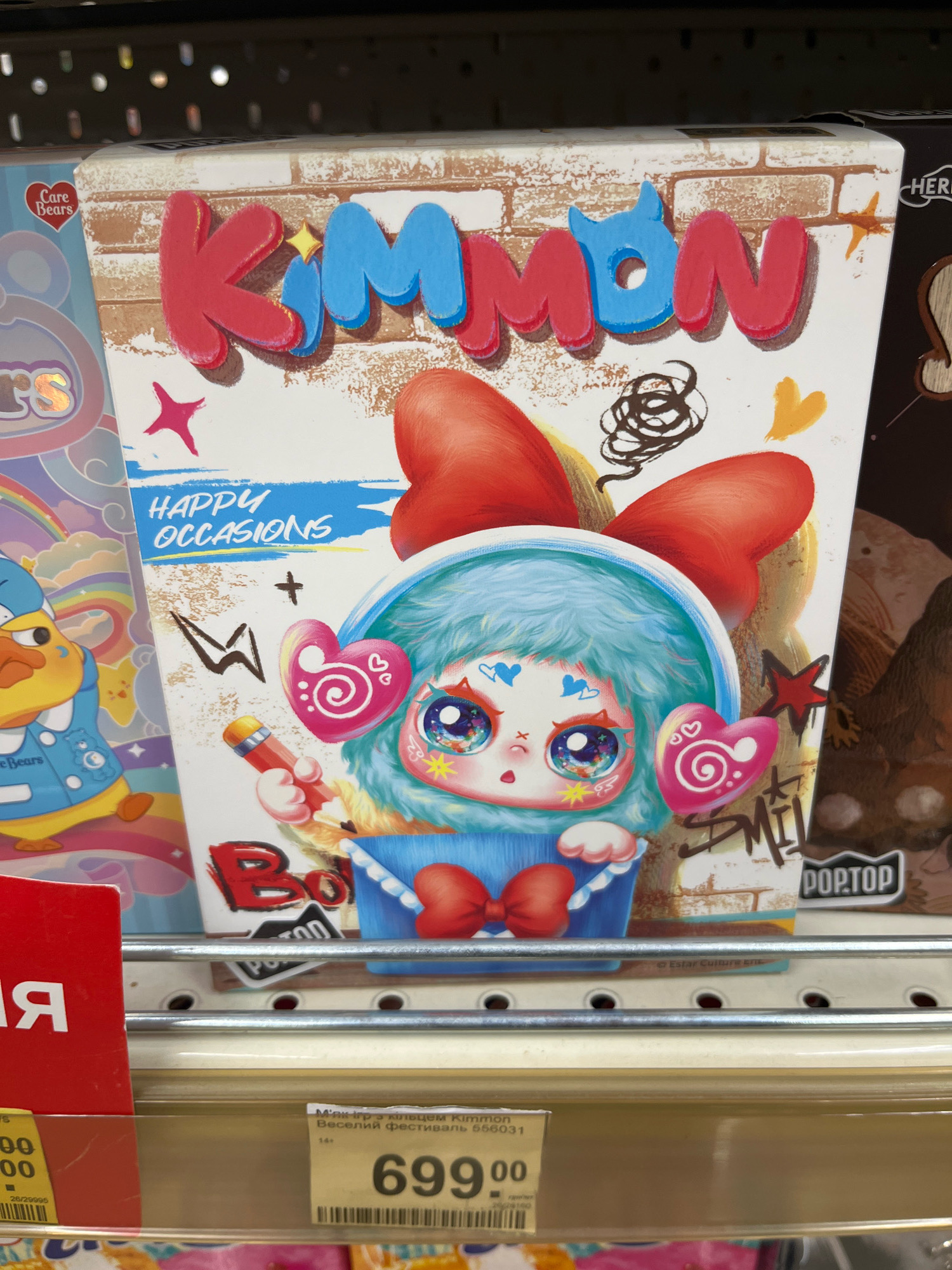 The image shows a close-up of a toy box on a shelf in a retail store. The toy box features a colorful illustration of a character with large eyes and fluffy blue hair, wearing a blue outfit and a red bow. The brand name "KIMMON" is prominently displayed in large, red and blue lettering at the top of the box. Below the character, the words "HAPPY OCCASIONS" are written. To the left of the KIMMON box, another toy packaging with a rainbow and a duck character is partially visible. To the right, a brown box with the text "POP.TOP" is also visible. The shelf is made of metal, and the background suggests a retail environment with other products on shelves above. A price tag with the number "699" is visible below the KIMMON box, along with some Cyrillic text. The overall scene is well-lit and appears to be a product display in a toy store. There is no visible indication of the city of Kharkiv, Ukraine, in the image itself.