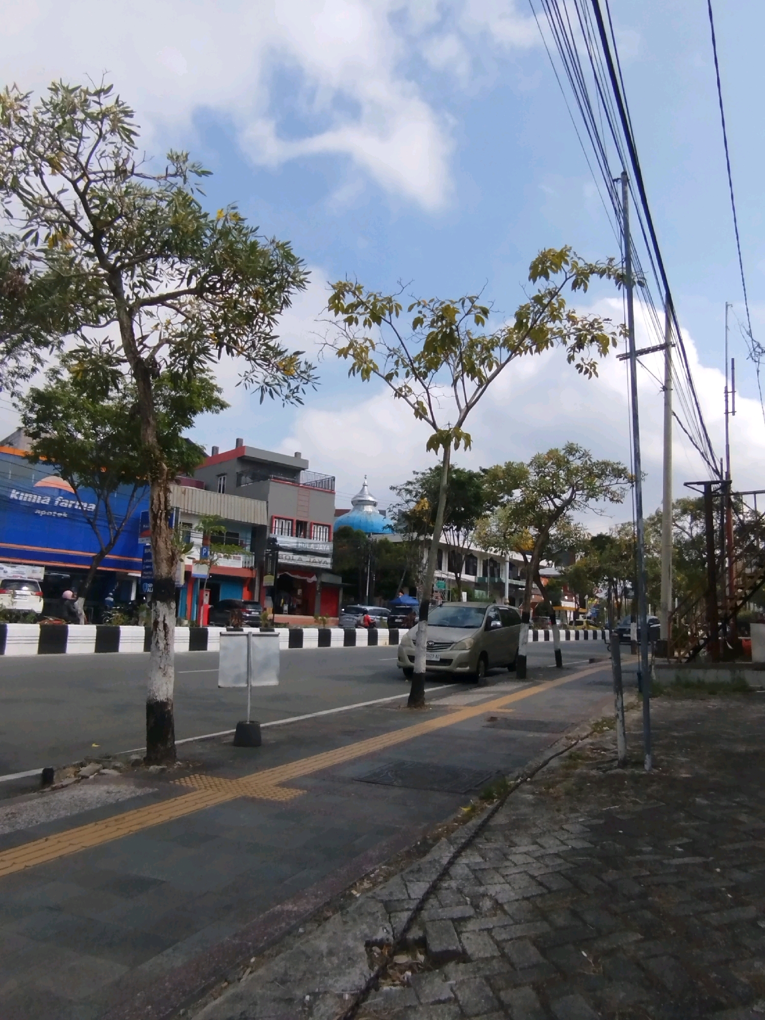 The image shows a street scene in Balikpapan, Indonesia. On the left side of the road, there are several buildings, including a large blue building with the text "Kimia Farma apotek" displayed prominently. Next to it, there are other buildings with various architectural styles and signage. A gold-colored car is driving on the road in the center of the image. Several trees are planted along the sidewalk and street. Overhead, a network of power lines runs across the sky, which is blue with scattered white clouds. The sidewalk is paved with dark tiles and has a yellow tactile paving strip. A metal pole with signage is visible on the sidewalk. The road is marked with black and white diagonal stripes along its edge.