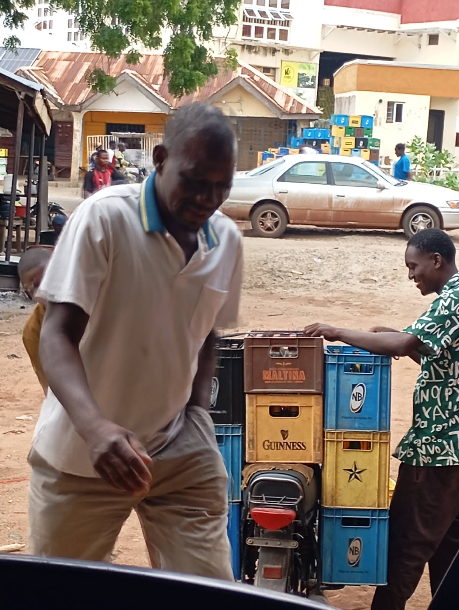 A medium shot captures an outdoor scene in Shendam, Nigeria, where a Black man, appearing to be in his late 30s or early 40s, stands prominently in the foreground. He is wearing a light-colored polo shirt with a blue and yellow collar and light brown trousers, his left hand is in his pocket, and he looks down and to his right. Behind him, stacked crates containing beverages are visible, with some labeled "MALTINA" and "GUINNESS." To the right of the crates, another Black man in a green patterned shirt is visible, smiling and reaching towards the crates. In the background, a silver sedan is parked, and further back, buildings with corrugated metal roofs and some vegetation are present. The ground is covered in dirt and sand. A portion of a motorcycle is visible below the crates. The overall setting suggests a commercial or informal marketplace environment.