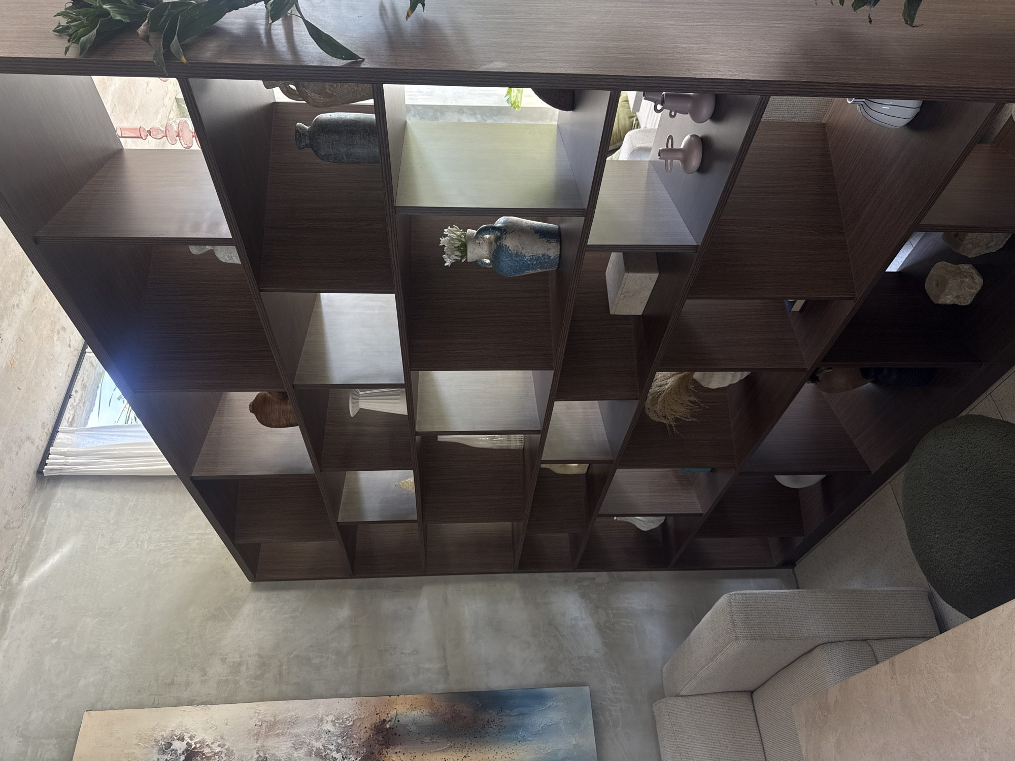 A bookshelf with various objects sits in a room in Ubud District, Indonesia. The objects include vases, rocks, and a few decorative items. The room is decorated in shades of gray, with a sofa and round chair nearby. A painting hangs on the wall below the bookshelf. Natural light comes through a window in the background. The bookshelf appears to be the focal point of the room.