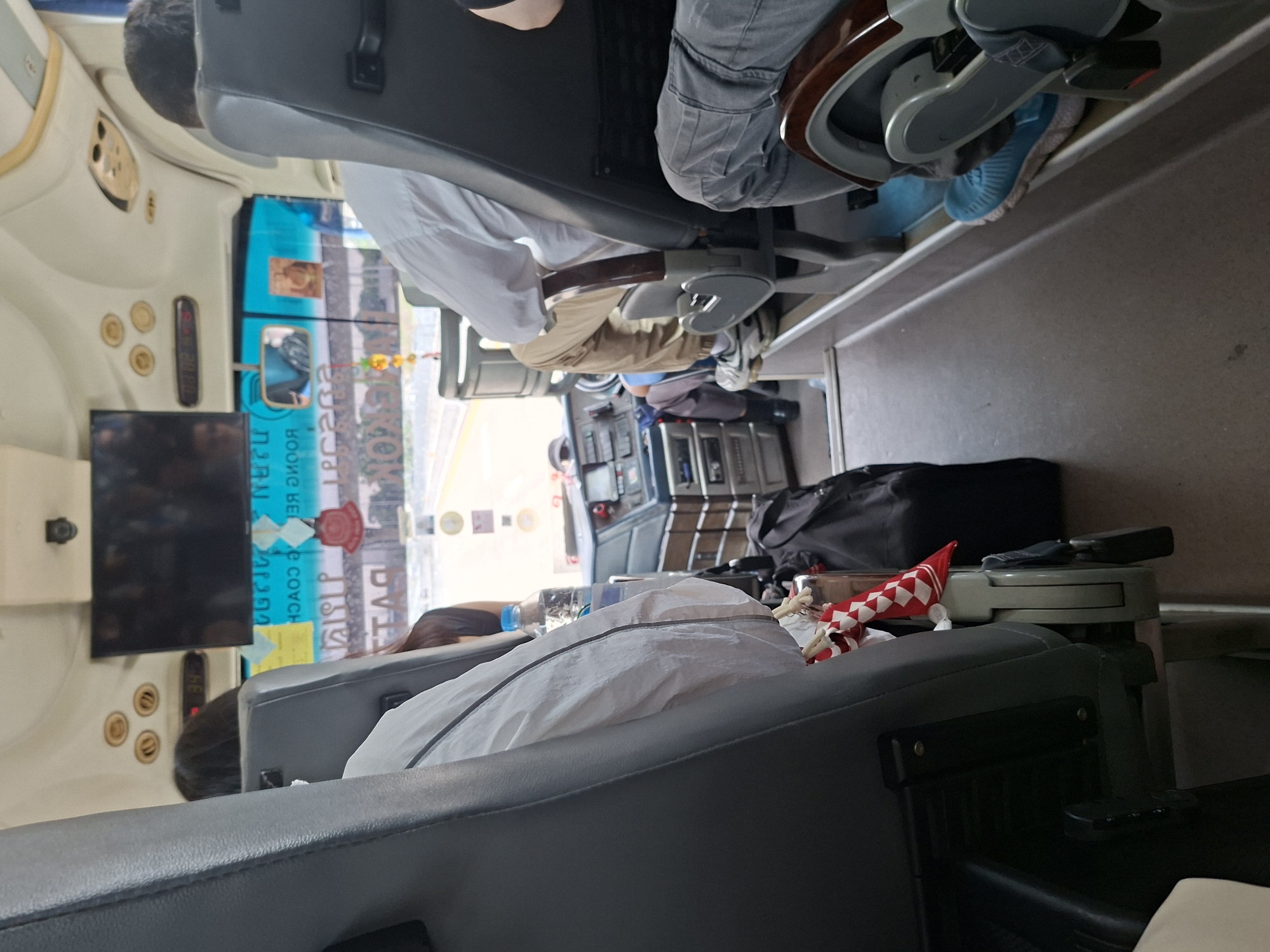 The image shows the interior of a vehicle in บ้านหนองน้ำเต้าลอย, Thailand. Passengers are seated in gray seats. A television screen is visible, and there is luggage in the aisle. One passenger wears blue shoes. A table contains a water bottle and an object with red and white stripes.