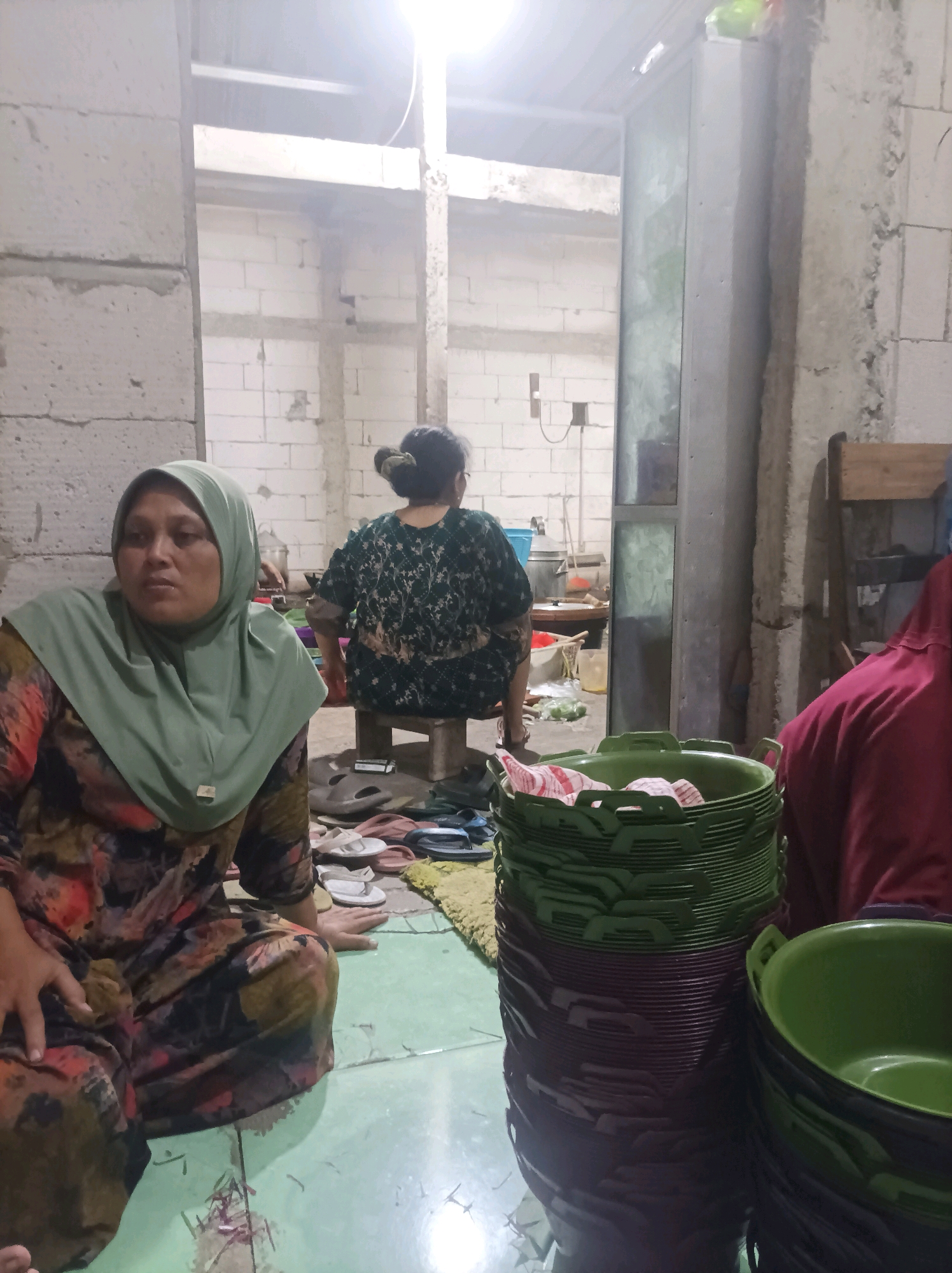 In Canga'an, Indonesia, two women are present in an unfinished room with brick walls. One woman, wearing a green hijab and a patterned dress, sits on the floor, looking to her left with her hands in her lap. Another woman, facing away from the camera, is seated on a low wooden stool, wearing a dark green patterned shirt. In the foreground, to the right of the seated woman, a large stack of green plastic bowls and a few other items are visible. The room appears to be under construction or renovation, with exposed brick and concrete elements. The lighting is bright, suggesting artificial light sources.