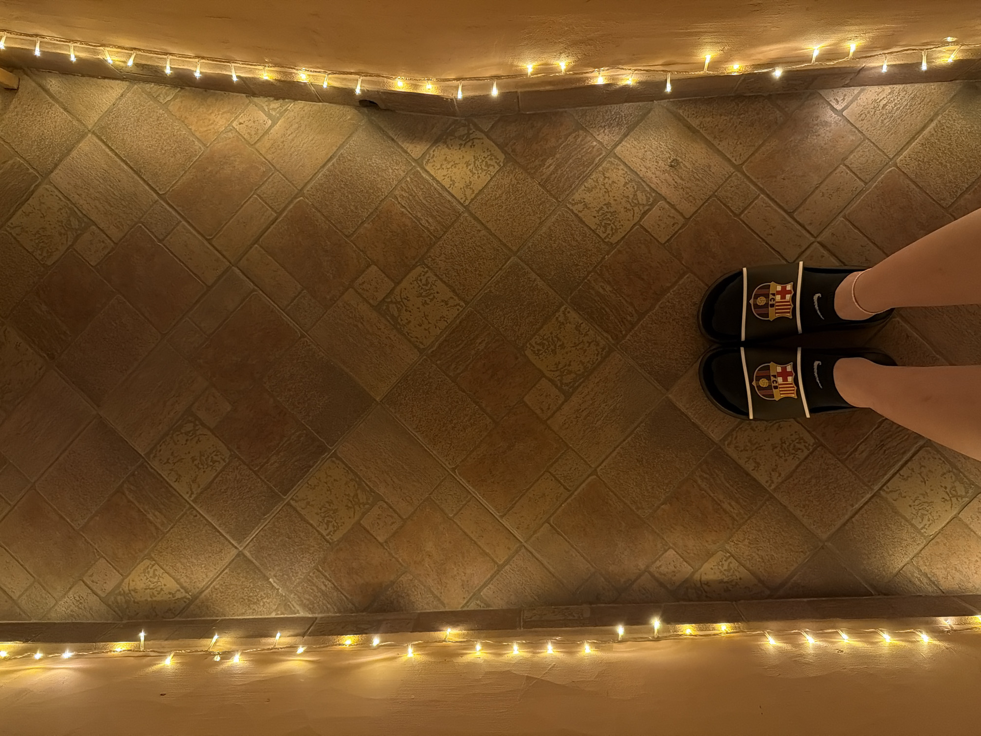 The image captures a top-down view of a person's legs and feet standing on a tiled floor in an indoor setting, likely a hallway or pathway, in Lovech, Bulgaria. The floor consists of light brown and beige tiles arranged in a diagonal pattern of squares and rectangles, some with subtle variations in texture and shade.
The subject is wearing black slide sandals, one on each foot. Each sandal strap prominently displays the colorful crest of the FC Barcelona football club and a white Nike swoosh logo. A delicate, light-colored anklet adorns the right ankle.
The scene is softly illuminated by two strings of warm, glowing fairy lights. One string runs along the upper edge of the frame, and another along the lower edge, creating a decorative and cozy ambiance. These lights cast gentle reflections on the tiled floor and illuminate the nearby light-colored wall or ledge surfaces to which they are attached. The presence of these lights suggests either an evening or nighttime setting, or an otherwise dimly lit indoor environment.
No specific activity beyond standing is depicted. The visible text elements are the "Nike" swoosh and the "FC Barcelona" crest on the sandals.