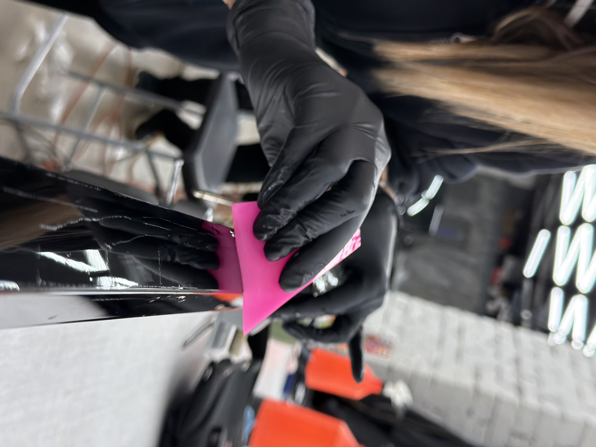 A pair of hands wearing black latex gloves are using a pink squeegee to apply something to a glossy black surface. The application appears to be precise and meticulous. Behind the hands and surface are indistinct objects and lighting, possibly within a work environment. Based on the surroundings, the location may be Krasnoyarsk, Russia. However, lacking more definitive markers or context, the geographical location is uncertain.