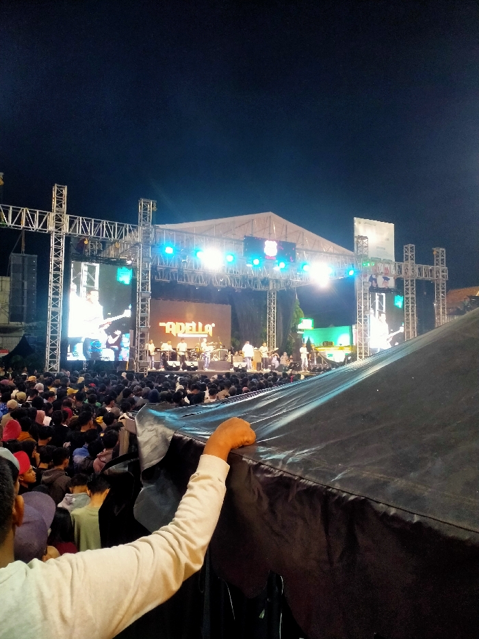 The image shows a large crowd gathered for a concert, possibly in a park or outdoor venue, at night in Boyolali, Indonesia. The stage is decorated with lights and banners. A large screen displays an image of a band performing. Many people are present, facing the stage. The image is taken from the perspective of a person seated in the crowd, looking towards the stage. The person's arm is seen reaching out towards the camera, touching the edge of a black tent that is positioned in front of the viewer. The image captures the energy and excitement of a live concert in Boyolali, Indonesia.