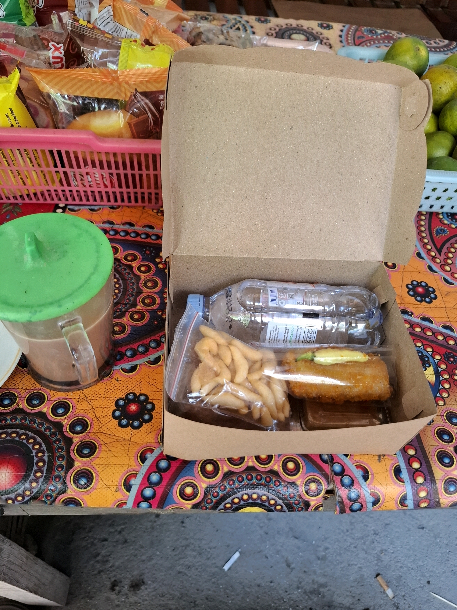 In Kedungmaron, Indonesia, a close-up, high-angle shot captures a takeout meal box on a vibrantly patterned tablecloth. The meal box, made of brown cardboard, is open, revealing its contents. Inside, a plastic-wrapped portion of french fries sits next to a breaded, fried item topped with a small green chili and yellow sauce. A clear plastic bottle of water is positioned behind the food. To the left of the meal box, a transparent mug with a green lid contains a brown liquid, likely coffee or tea. A pink plastic basket filled with various snack packages is visible behind the mug. The background includes out-of-focus elements like more snack packages, a basket of green oranges, and a glimpse of what appears to be a wooden structure. The ground in the foreground is concrete, with several cigarette butts scattered on it.