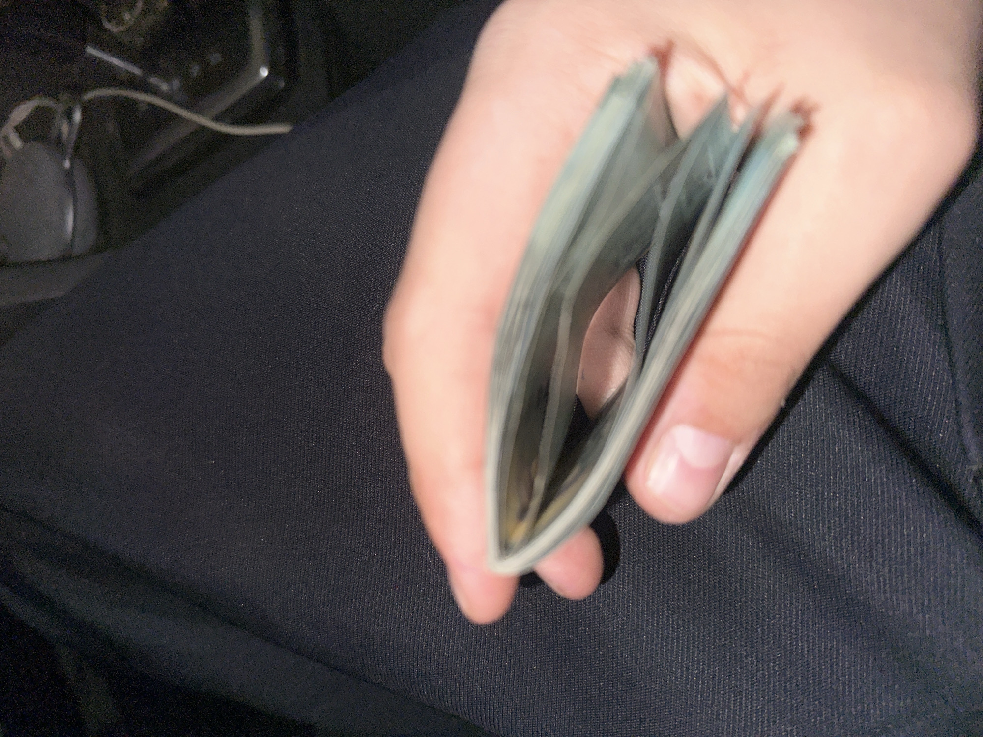 A person's hand is holding a stack of cash, appearing to be US dollar bills. The background shows a portion of a dark-colored lap, possibly pants, and a blurred object that could be a computer mouse. The location is unknown.