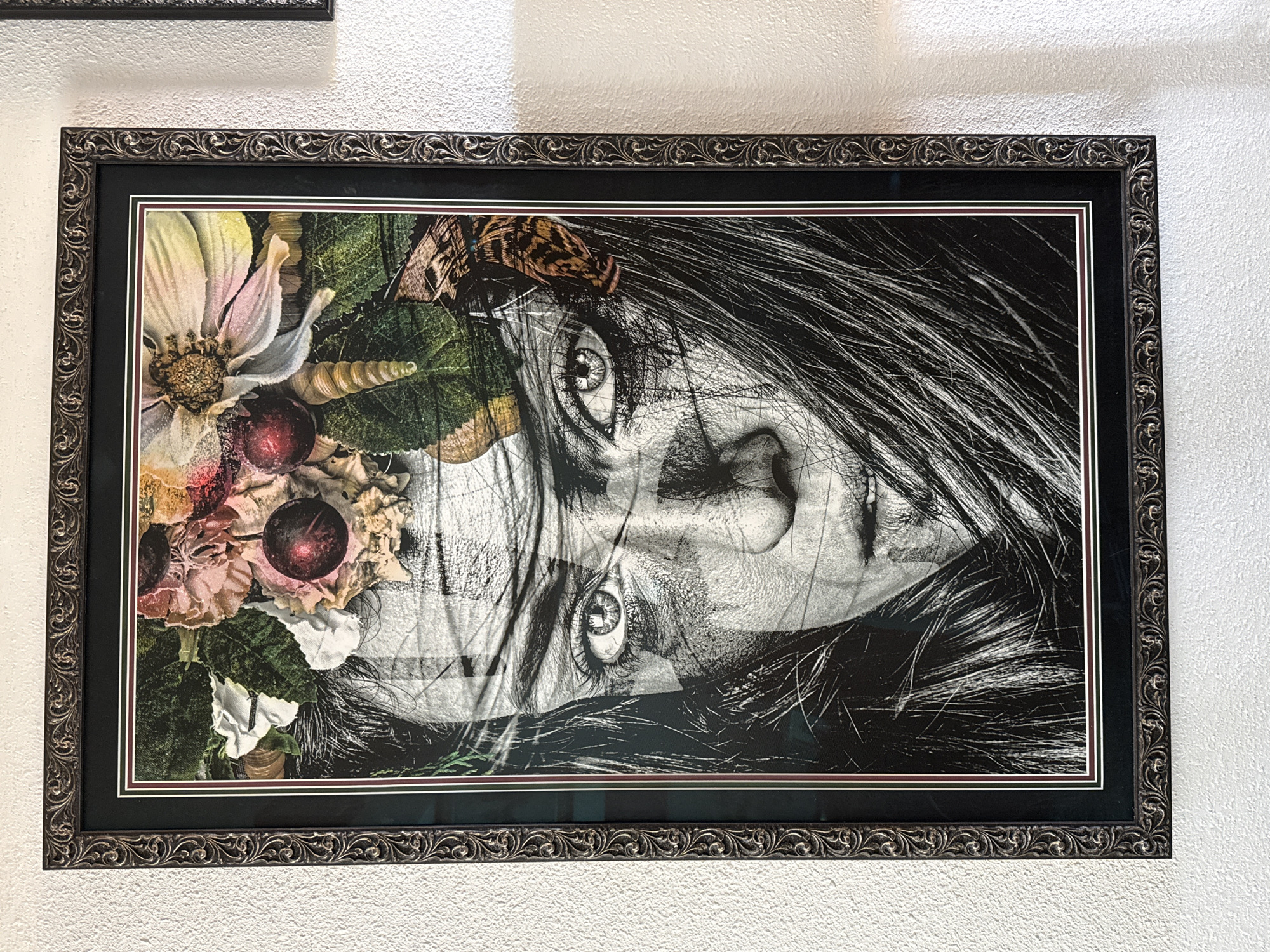 The image displays a framed artwork hanging on a textured white wall. The artwork is a close-up, black and white portrait of a person's face, partially obscured by flowing dark hair. The subject's eyes are prominently featured, appearing wide and intense. Elements of nature, including flowers, cherries, leaves, a seashell, and a butterfly, are artfully arranged around the subject's head as if forming a headdress. The frame is ornate and dark, with intricate scrollwork. The lighting suggests an indoor setting, possibly a home or gallery. The overall style of the artwork is a blend of realism and surrealism. The location in Tacoma, United States of America, is not discernible from the image itself.