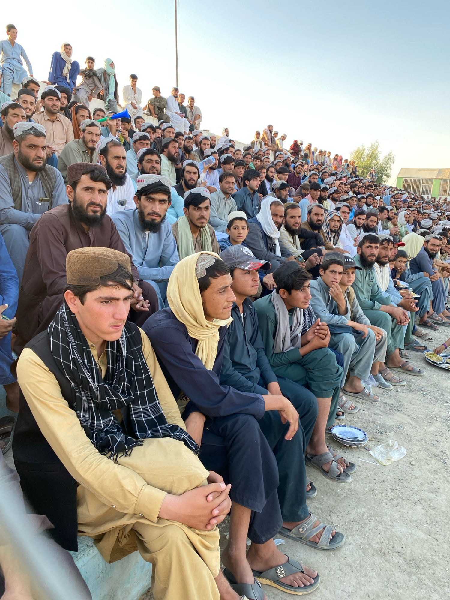 The photograph captures a large gathering of men and boys sitting in tiered seating, likely a stadium or outdoor venue, under a clear sky. The setting appears to be in Spin Boldak, Afghanistan, with a low-lying building or structure visible in the background. The individuals are predominantly dressed in traditional Afghan clothing, including shalwar kameez and embroidered caps (pakols). Many of the men have beards. The seating is a rough, concrete structure. In the foreground, a young man in a yellow shalwar kameez, black vest, and a black and white checkered scarf is prominently seated. Some individuals are looking towards the right of the frame, suggesting an event or performance is taking place. Plates and discarded plastic are visible on the ground in the lower right quadrant, indicating that food may have been consumed or is being served. The overall atmosphere seems to be one of a community event or gathering.