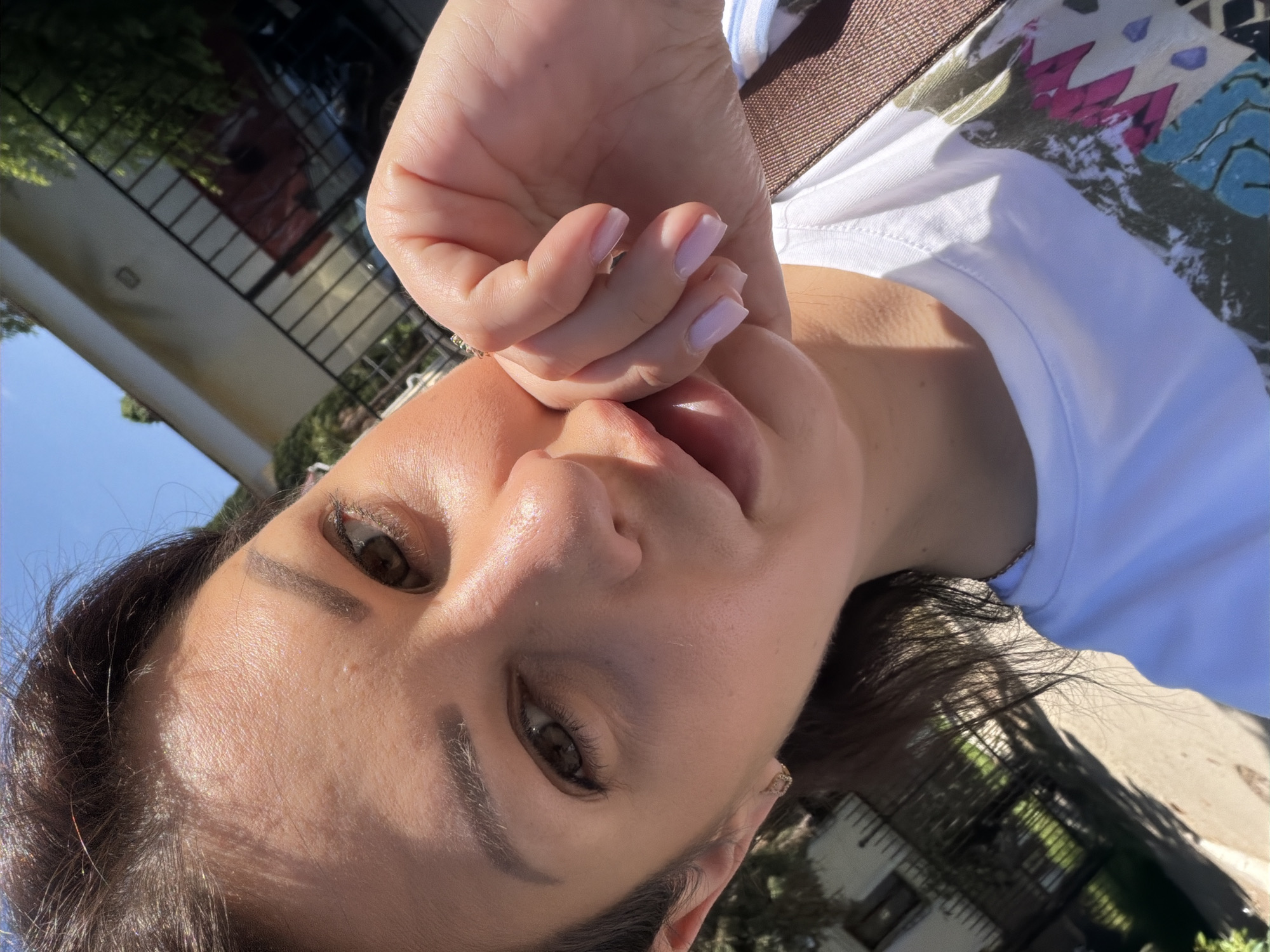 This image is a close-up, outdoor selfie of a female individual taken in the city of Sochi, Russia. The subject, with dark brown hair and brown eyes, is looking slightly downwards towards the camera. Her right hand, featuring light pink manicured fingernails, is positioned with fingers curled near her chin and lower lip. She wears a white t-shirt with a multi-colored abstract graphic design on the lower portion, incorporating green, pink, blue, and black elements. A small, gold-toned square or hexagonal earring is visible in her left ear. The scene is illuminated by strong direct sunlight, creating pronounced highlights on her face and hair. The background consists of a clear blue sky, a light-colored building, a dark metal fence or railing, and green trees and foliage, indicating a residential or suburban outdoor setting.