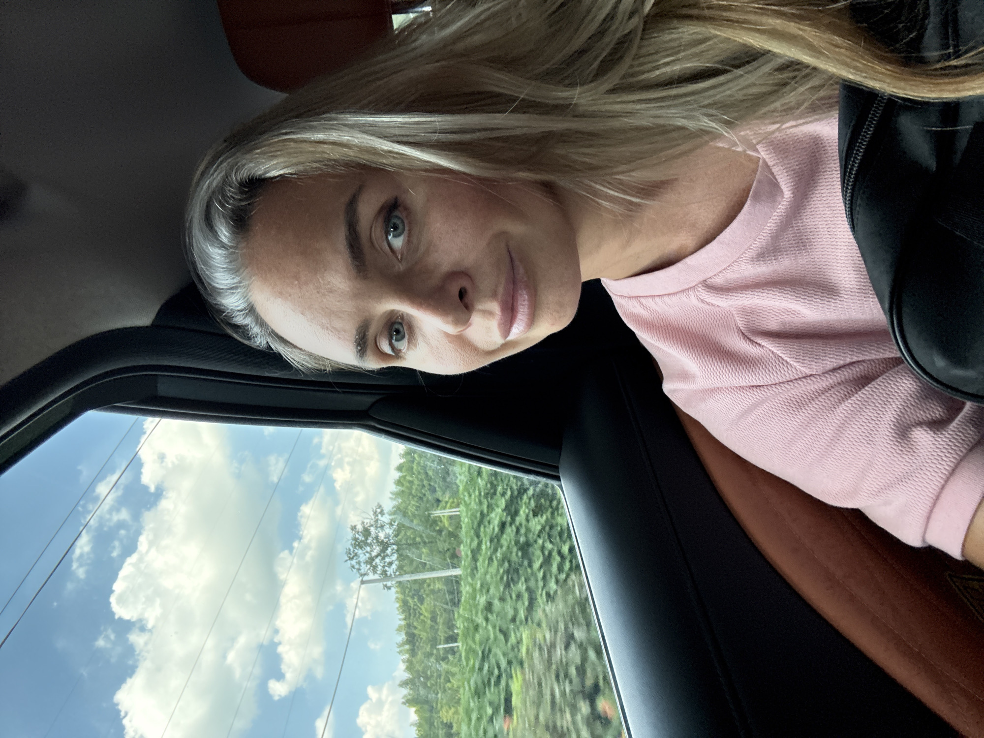 A woman with blonde hair and blue eyes is sitting in the back seat of a car, looking at the camera and smiling. She is wearing a pink shirt. Outside the car window, a green landscape with trees and power lines is visible under a blue sky with white clouds. The car's interior, including a brown leather seat and black trim, is also visible. The image appears to be taken in Wuxing District, China, given the landscape and greenery.