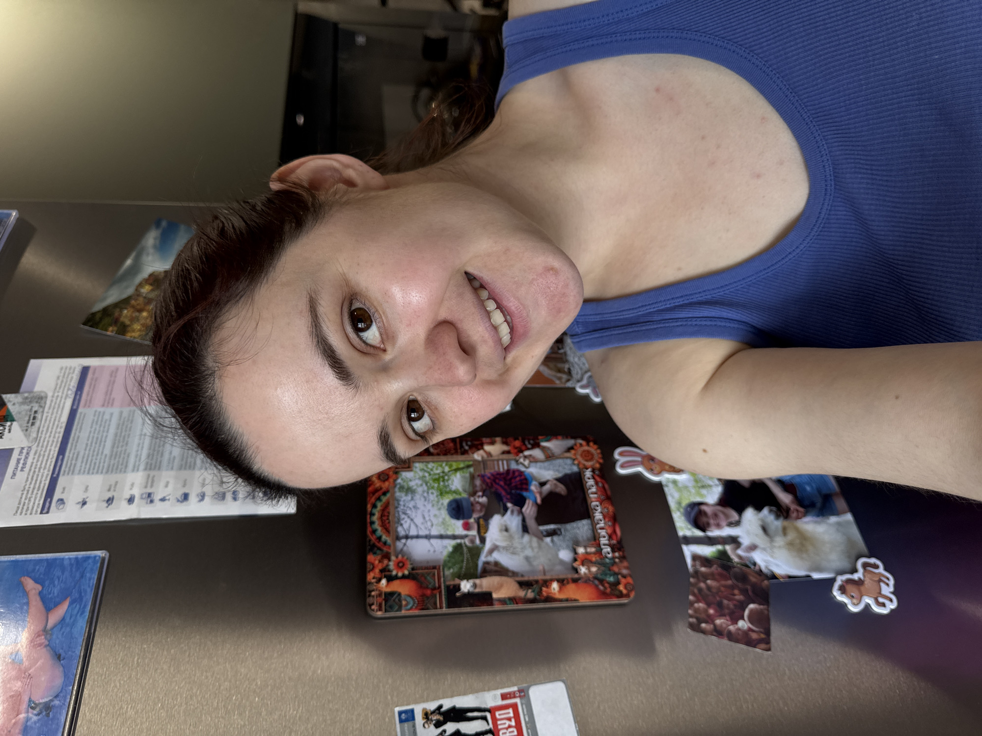 A woman with dark hair is posing in front of a refrigerator. The refrigerator has several magnets and pictures attached. One of the picture magnets appears to be of a family with a child and a Samoyed dog. Another magnet seems to feature an illustration of a cowboy. The text "IZH" is partially visible on one of the magnets, suggesting the city of Izhevsk, Russia. The woman is wearing a blue tank top and smiling.