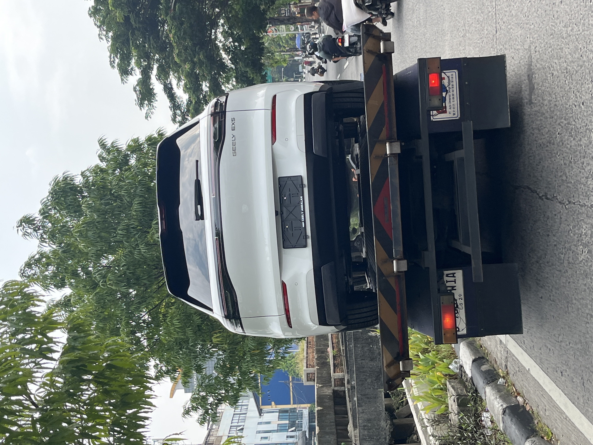 A white Geely EX5 SUV is prominently featured, loaded onto the back of a flatbed tow truck and facing away from the viewer. The tow truck has distinctive yellow and black diagonal warning stripes on its rear support structure, with red tail lights illuminated.
The scene depicts an urban setting, likely during the daytime under an overcast sky. On the road behind the tow truck, several motorcycles are visible, with some people riding them and others standing or crouching beside them. Lush green trees line the street, with a mix of buildings and structures in the background, including one with a distinctive blue facade. A concrete curb separates the road from a small strip of vegetation.
Visible text on the SUV's rear includes "GEELY EX5" on the tailgate and "GEELY ANDALAN" on the license plate frame, though the actual license plate is blank. On the back panel of the tow truck, text for "PT. Astra Universal Satya" is visible, along with phone numbers (e.g., "021 639..."). A partial license plate on the tow truck reads "...WIA" and "08.29".