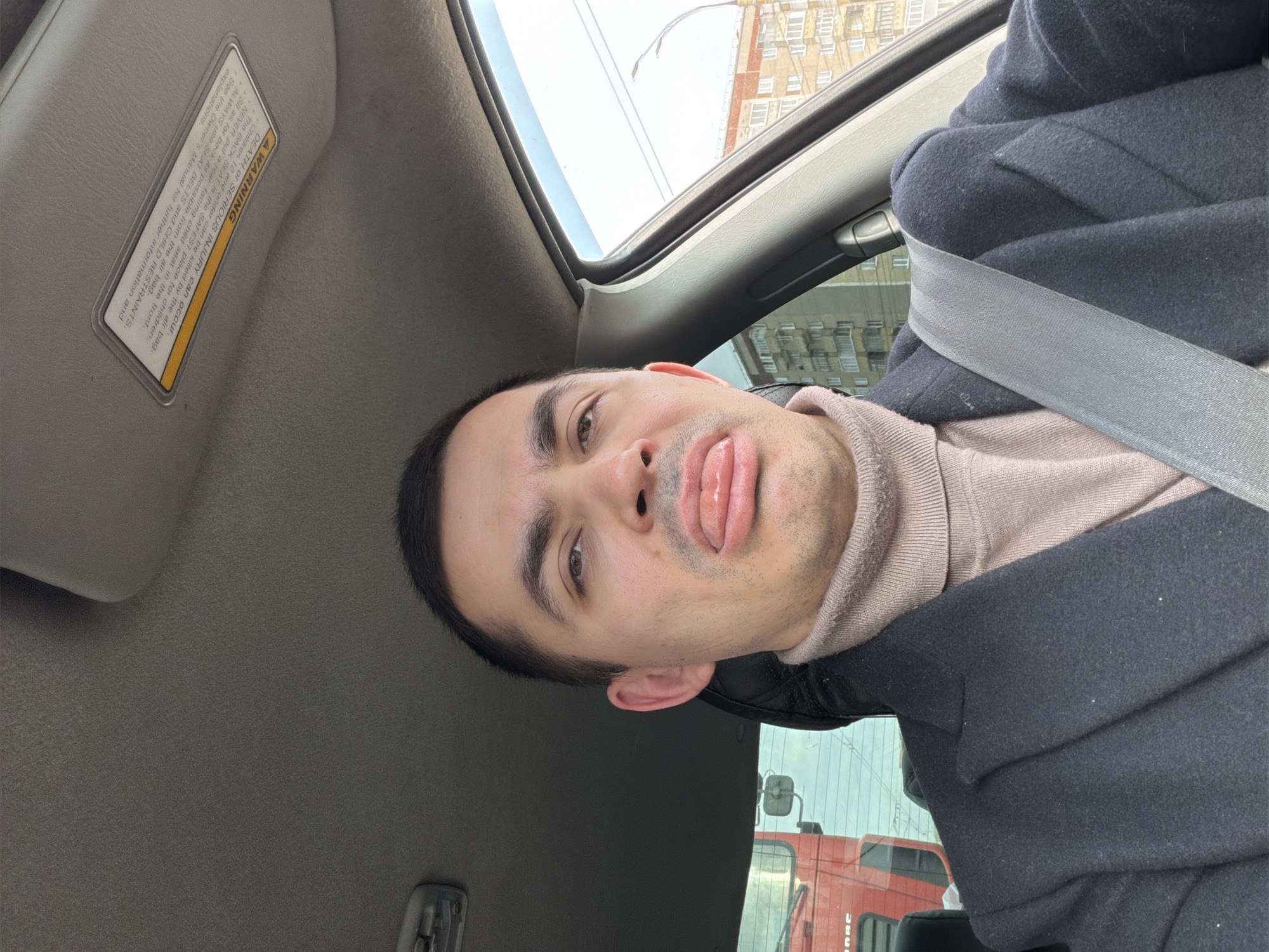 A man is taking a selfie inside a vehicle. He is wearing a black coat over a light pink turtleneck sweater and has a seatbelt buckled across his chest. The car's sun visor is visible, as well as the vehicle's interior. Outside the car window, buildings are visible, as is a red truck. Based on the building and the truck, this photograph was most likely taken in Izhevsk, Russia. The man is sticking his tongue out.