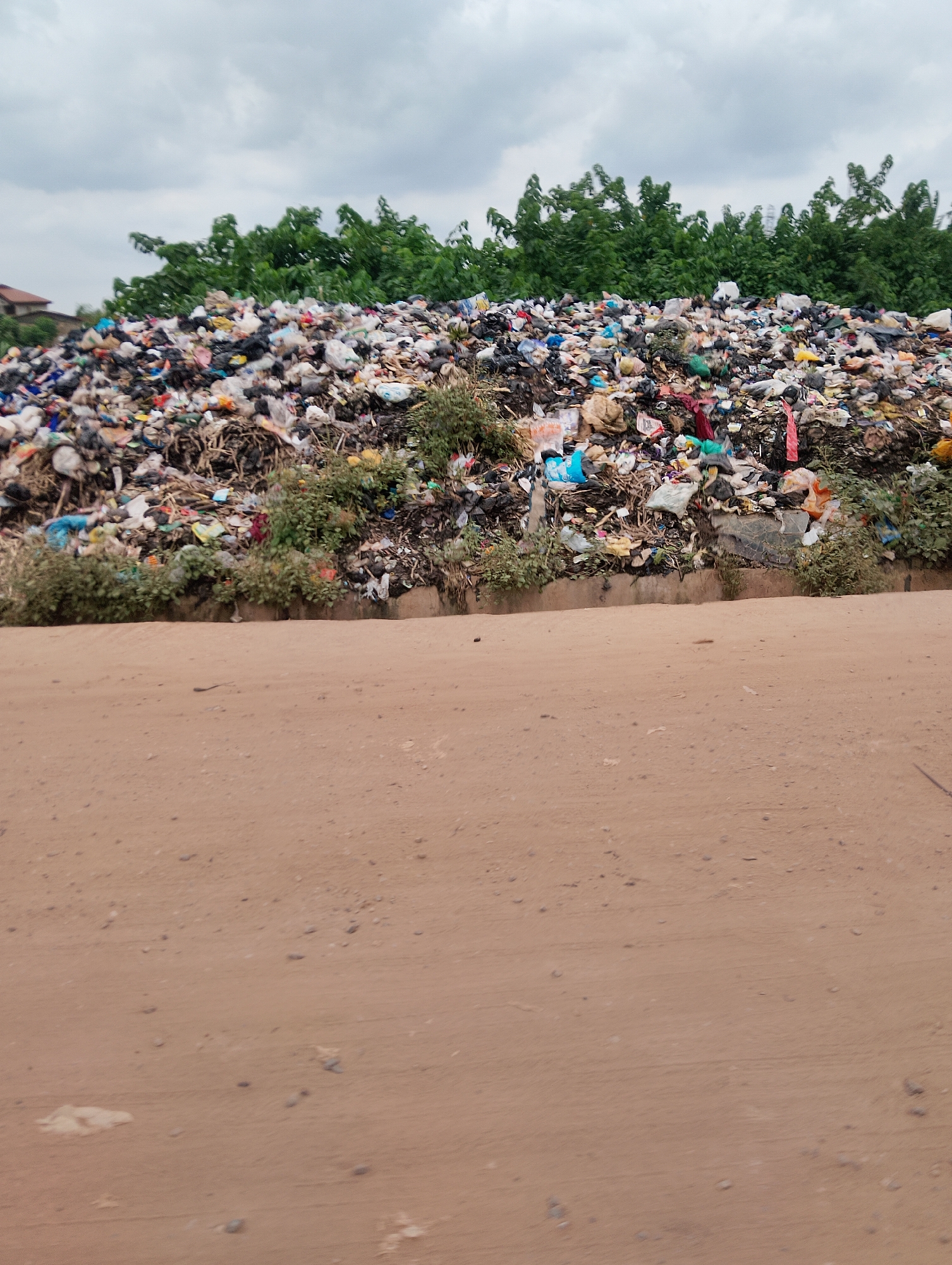 The image depicts a large, overflowing landfill located in Ijoko, Nigeria. The landfill is a massive mound of refuse, composed of various types of trash, including plastic bags, bottles, and organic waste. Lush green trees form a backdrop to the immense pile of garbage, and a cloudy sky hangs overhead. In the foreground, a sandy dirt road separates the viewer from the landfill. The scene is one of significant environmental neglect, with the sheer volume of waste creating an unsightly and potentially hazardous landscape.