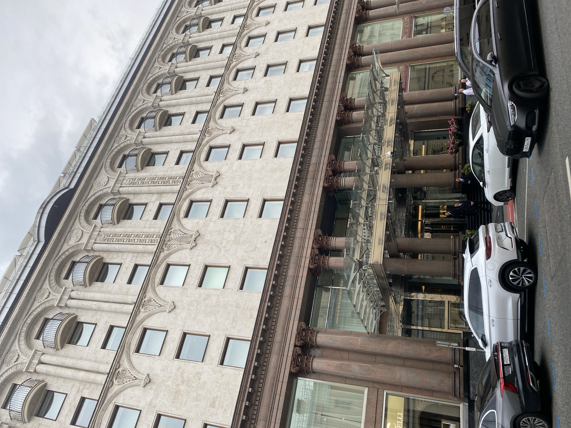 The image shows the grand facade of a historic building, likely a hotel, in Moscow, Russia. The building features ornate architectural details, including arched windows, decorative balconies with wrought-iron railings, and columns supporting a glass canopy over the entrance. Several cars are parked on the street in front of the building, including a black sedan and a white SUV. People can be seen entering and exiting the building. The sky is overcast, suggesting a cloudy day. The overall scene conveys a sense of elegance and grandeur, characteristic of Moscow's historic cityscape.