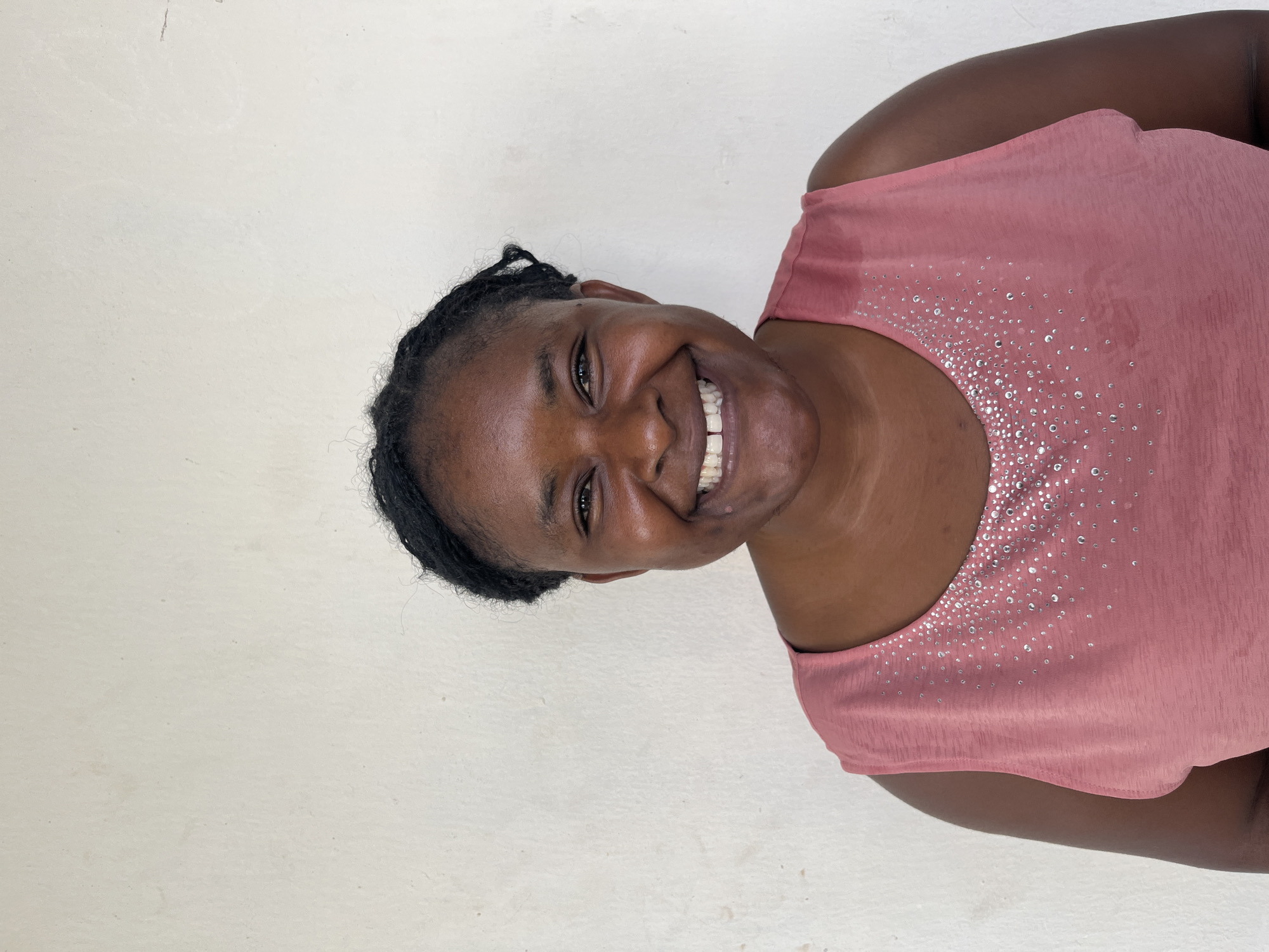 A head-and-shoulders studio portrait of a Black woman with dark skin and braided hair, taken against a plain, off-white wall. She is smiling broadly, her teeth showing, and her head is tilted slightly to her right. She is wearing a sleeveless, pink top with a scoop neck. The top is adorned with many small, sparkling rhinestones clustered around the neckline. The background is a textured, off-white wall with some faint dark smudges. The lighting is bright, creating a slight sheen on her forehead and chest. The image appears to have been taken outdoors or in a well-lit interior space.