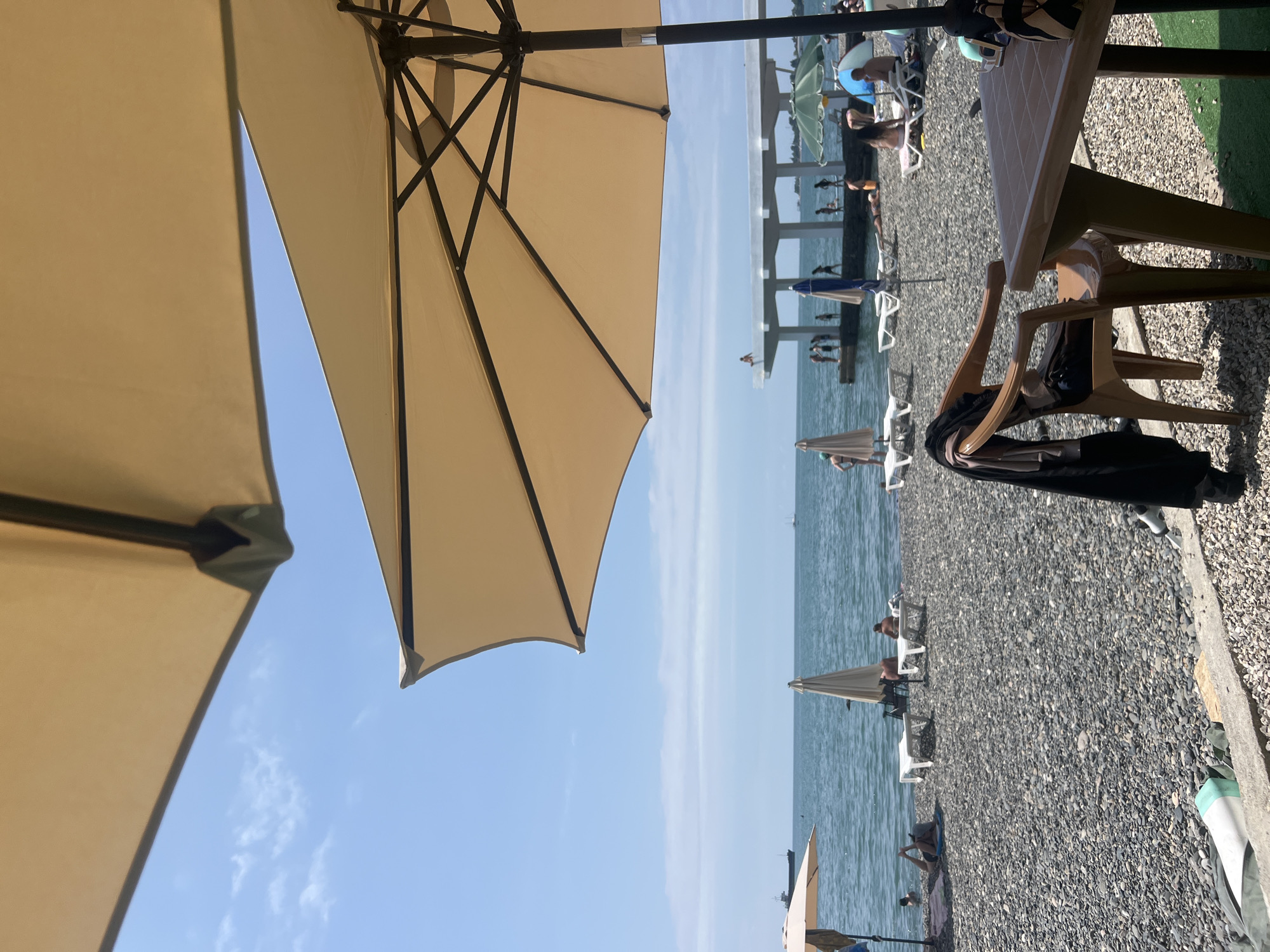 The image depicts a pebbled beach on a sunny day, likely in Sokhumi, Abkhazia, given the presence of a distinctive white pier structure in the background. The foreground is dominated by two large, light-colored umbrellas, one of which is partially obscuring the view of the sky. Underneath one umbrella, a brown plastic chair sits at a small table. A dark garment is draped over the back of the chair. Beyond this seating area, the beach is populated with several white lounge chairs, some occupied by sunbathers. The sea is visible in the middle ground, with a large ship on the horizon. Further back, the white pier structure extends into the sea, with numerous figures visible on and around it. Several other umbrellas, including a green one, are scattered across the beach. The sky is a clear blue with sparse white clouds.