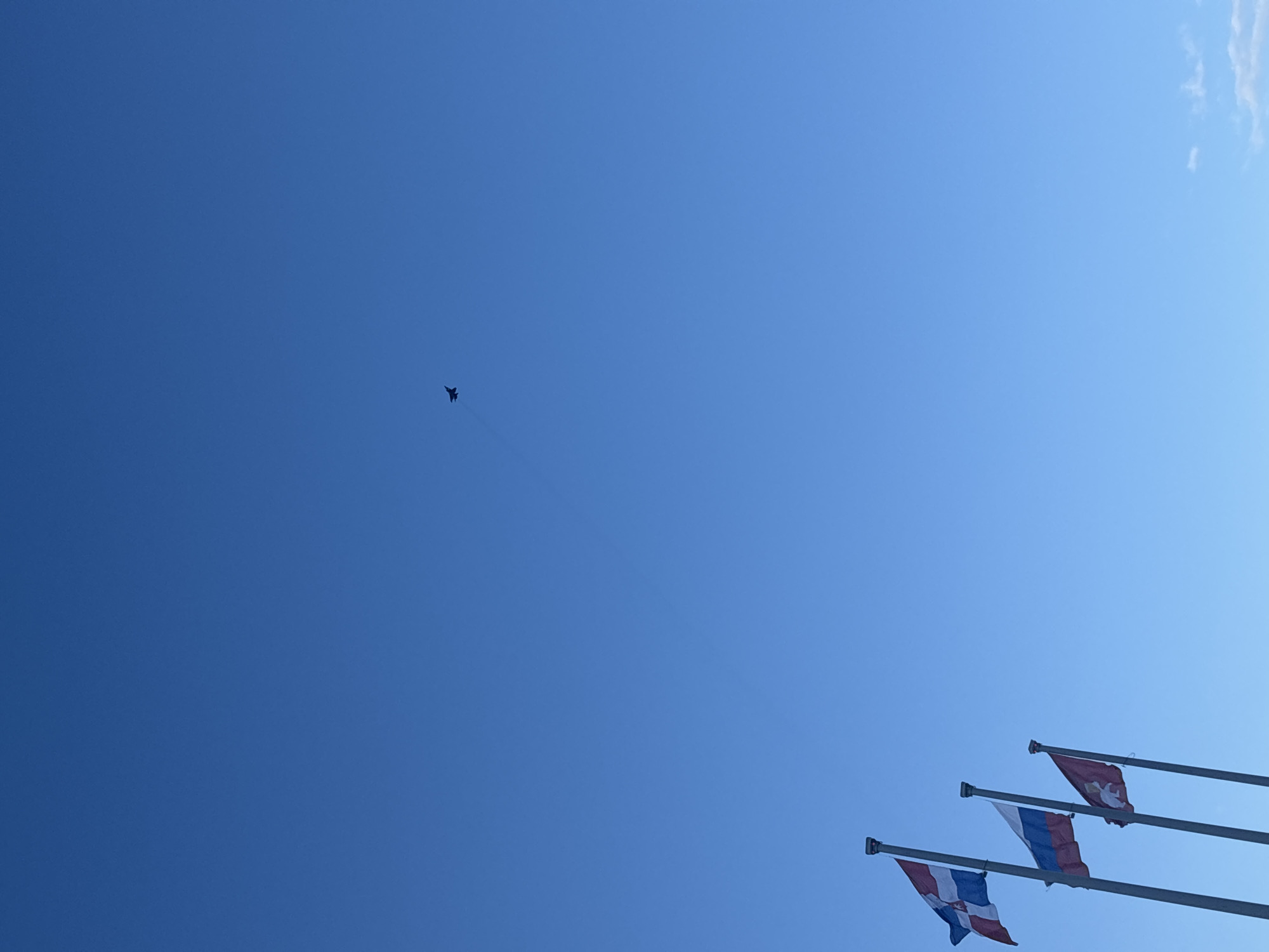 A bright, clear blue sky dominates the image, with a faint wisp of white cloud visible in the upper right corner. In the mid-left portion of the sky, a dark silhouette of a fighter jet, possibly a Mikoyan MiG-29 due to its distinct shape and twin tail fins, is captured mid-flight, leaving a subtle, thin contrail behind it.
In the bottom right corner, three flags are flying on flagpoles, partially obscured but mostly visible. From left to right:
1. The leftmost flag displays horizontal stripes of blue, white, and red (from top to bottom), with a dark emblem or coat of arms visible in its center. This configuration, often representing historical flags of the Perm Governorate or a variant of the Perm Krai flag, is shown alongside other official flags.
2. The middle flag is clearly the national flag of Russia, featuring horizontal stripes of white, blue, and red (from top to bottom).
3. The rightmost flag is predominantly red with a central emblem: a white, winged bear holding a golden Gospel book on its back and topped with a golden cross. This is the official flag of the City of Perm.
The scene takes place during daytime under sunny weather, indicated by the vibrant blue sky and distinct shadows. The combination of the Russian national flag and the specific flags of Perm Krai/City of Perm unequivocally places the location in Perm, Russia. No visible text can be clearly read from the flags or other elements in the image.