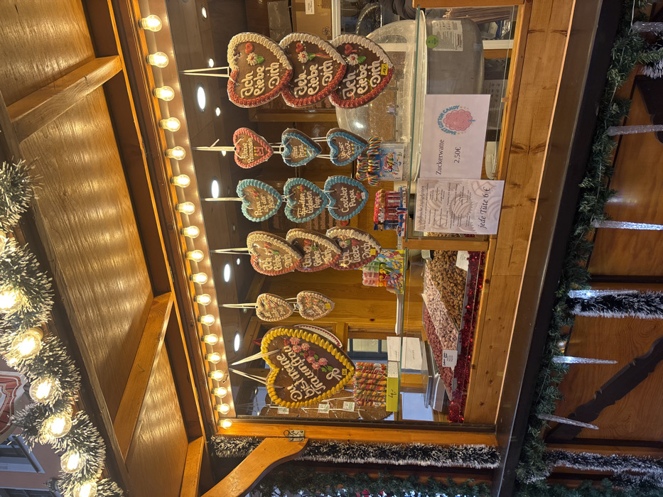 This image captures a festive wooden market stall, likely part of a Christmas market, situated in Meissen, Germany. The stall's exterior is decorated with green tinsel intertwined with warm white string lights along the roof's edge, and illuminated icicle ornaments hang from the lower counter. Inside the brightly lit display window, numerous heart-shaped gingerbread cookies (Lebkuchenherzen) are suspended. These gingerbread hearts feature various German phrases, including "Du bist meine Traumfrau" (You are my dream woman), "Ich liebe Dich" (I love you), "Papa ist der Beste" (Dad is the best), "Kleine Prinzessin" (Little Princess), "Unser Prinz" (Our Prince), "Frechdachs Papa" (Cheeky Dad), and "Coolster Papa" (Coolest Dad). One "Ich liebe Dich" heart also displays "ABV."
Below the hanging cookies, the counter presents several bins filled with various types of nuts or candied nuts, set on a bed of red tinsel. Colorful spiraled lollipops are visible on the left side of the counter, along with smaller packaged candies. White signs are displayed, one advertising "Zuckerwatte" (Cotton Candy) for "2,50€" with a graphic, and another indicating "jede Tüte 6€" (each bag 6€) for other items. A commercial scale is positioned on the left side of the counter, and a cotton candy machine is partially visible on the right. No individuals are present within the frame.