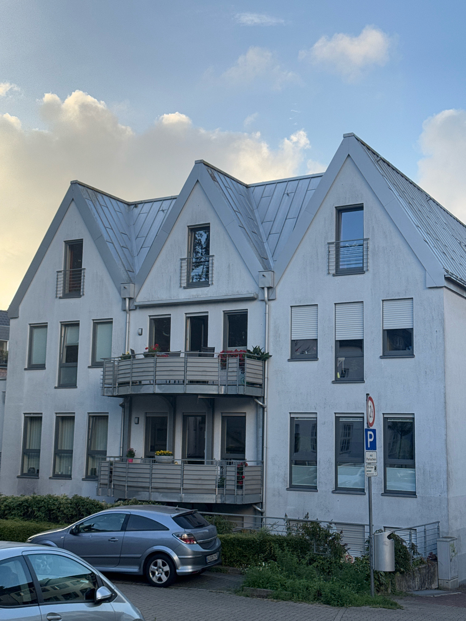 In the city of Cleves, Germany, a light gray, three-story apartment building with a metal pitched roof is shown. The building has multiple windows on each floor, some with blinds. Balconies with metal railings are present on the second and third floors. In front of the building, a light gray hatchback car is parked on a paved surface, with another car partially visible in the foreground. A signpost with a parking sign and weight limit information is located to the right of the building, near a trash can and overgrown bushes. The sky above is partly cloudy with a warm light suggesting either sunrise or sunset.