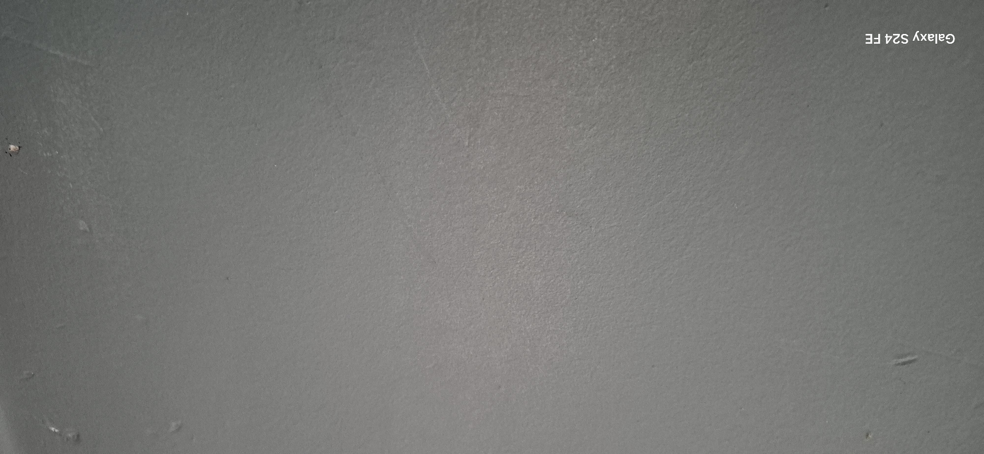 The image is a close-up, full-frame shot of a gray, textured surface, likely a wall or a floor. The texture appears slightly rough and mottled. There are a few small, subtle marks or imperfections visible on the surface. In the bottom left corner of the image, the text "Galaxy S24 FE" is present in white lettering, indicating the device used to capture the photograph. No people, specific objects, or discernible environmental context outside of the immediate surface are present. The lighting is even, with no strong shadows. The location is identified as Malko Tarnovo, Bulgaria, however, this information is not visually evident from the image itself.