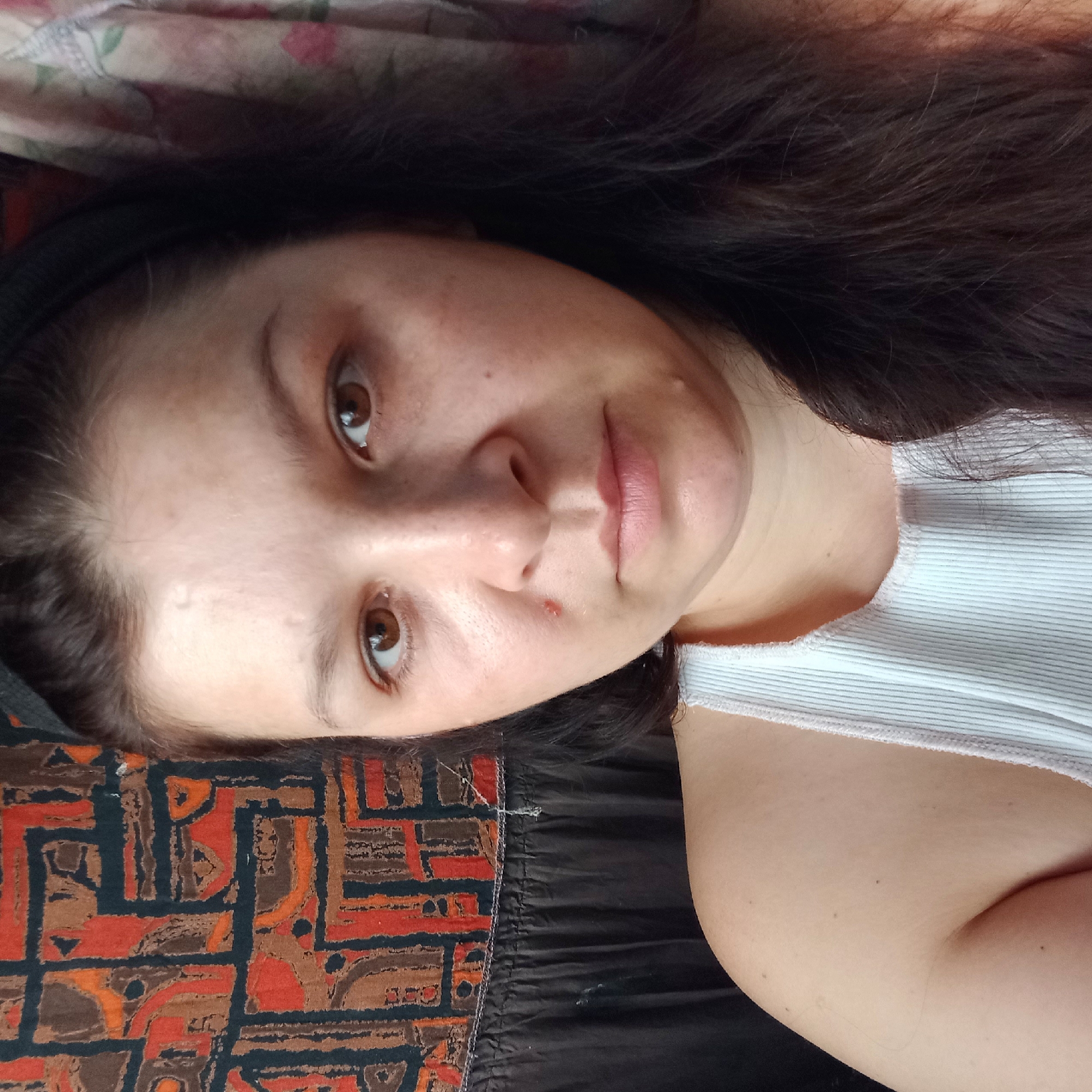 A close-up selfie of a woman with dark hair, brown eyes, and fair skin. She is wearing a white tank top and a black headband. Her face is the primary focus, with her head slightly tilted. A patterned fabric is visible behind her on the left, and a floral patterned fabric on the right. The scene appears to be indoors, likely a domestic setting. There is no information to indicate the location in the city of Klyntsi, Ukraine.