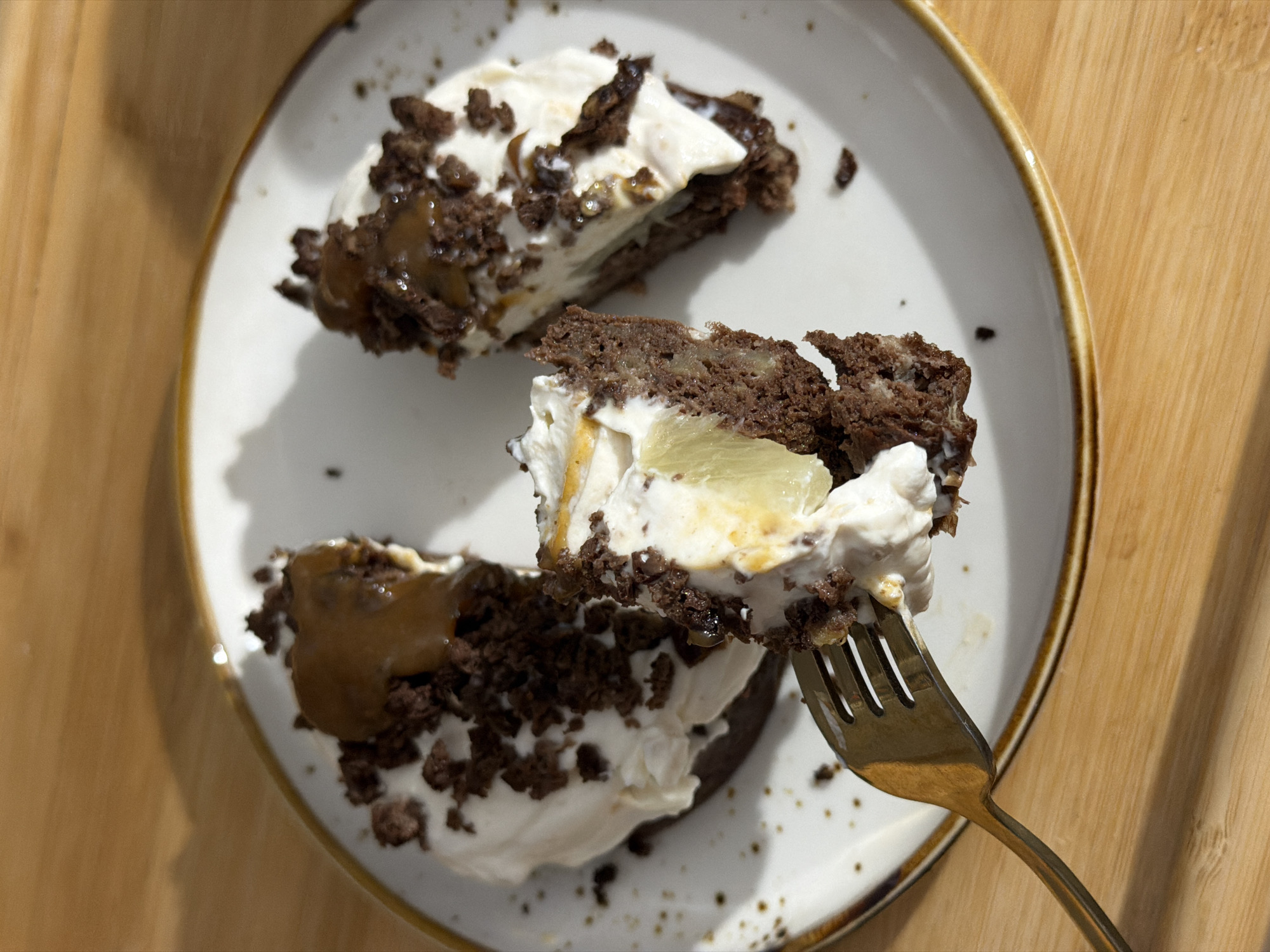 A partially consumed dessert, likely a cake or pastry, is displayed on a white ceramic plate with an irregular brown rim and sparse brown speckles. The dessert features a dark brown, crumbly base, topped with a thick layer of white cream. Scattered dark brown crumbs and a viscous, light brown liquid drizzle, consistent with caramel sauce, adorn the cream surface. A pale yellow, translucent fruit slice, possibly pineapple, is embedded within the cream and cake, visible in a removed section. A metallic fork, appearing to have a bronze or gold finish, holds a bite-sized piece of the dessert, lifting it from the plate. The dessert shows signs of active consumption, with multiple sections broken apart. The plate rests on a light brown wooden surface, illuminated by strong, directional lighting from the right, which casts distinct shadows. The image contains no visual information to determine its location within the city of Perm, Russia.